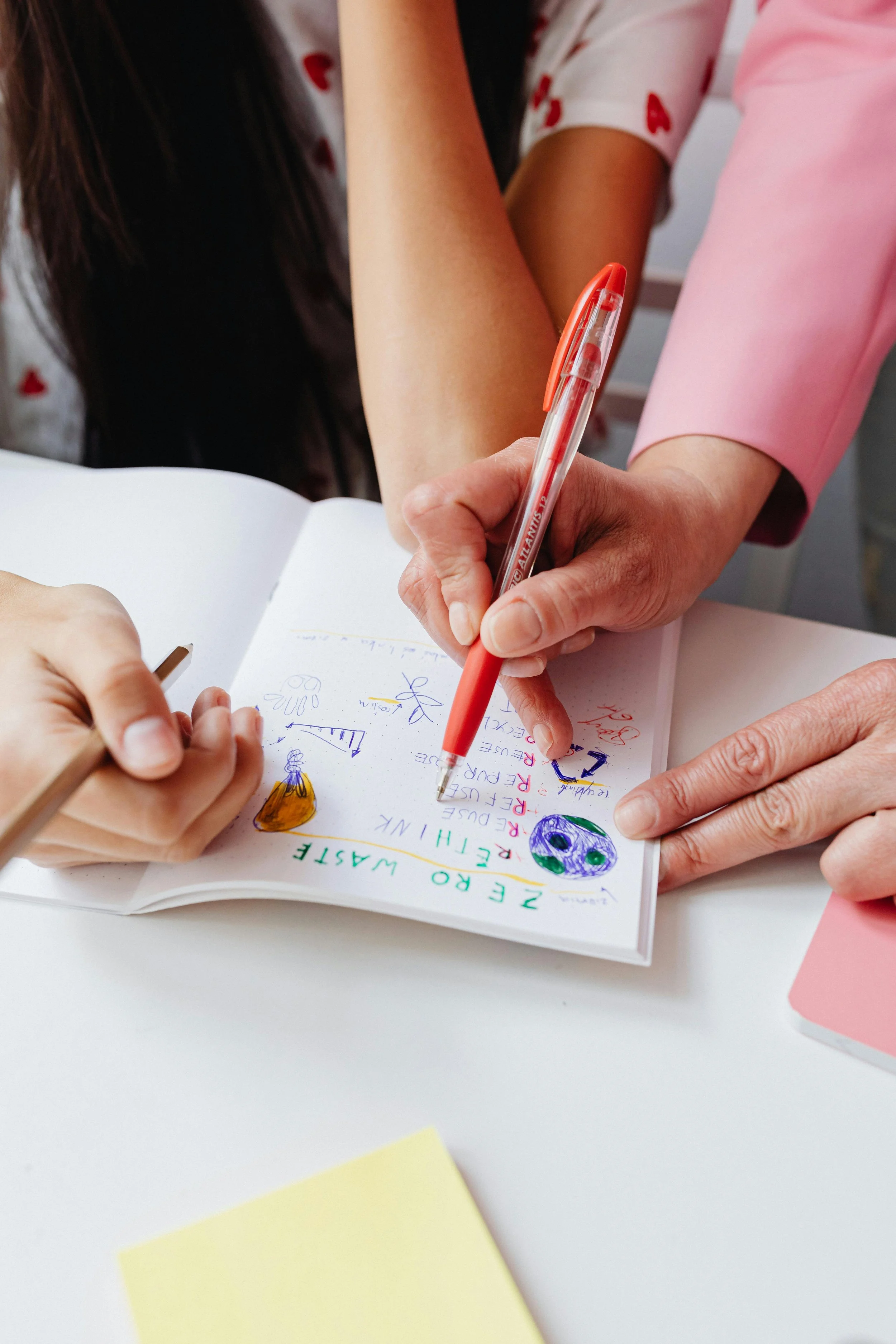 Two people working on a creative project, one holding a red pen and pointing at a colorful sketchbook, sketching and writing on it.
