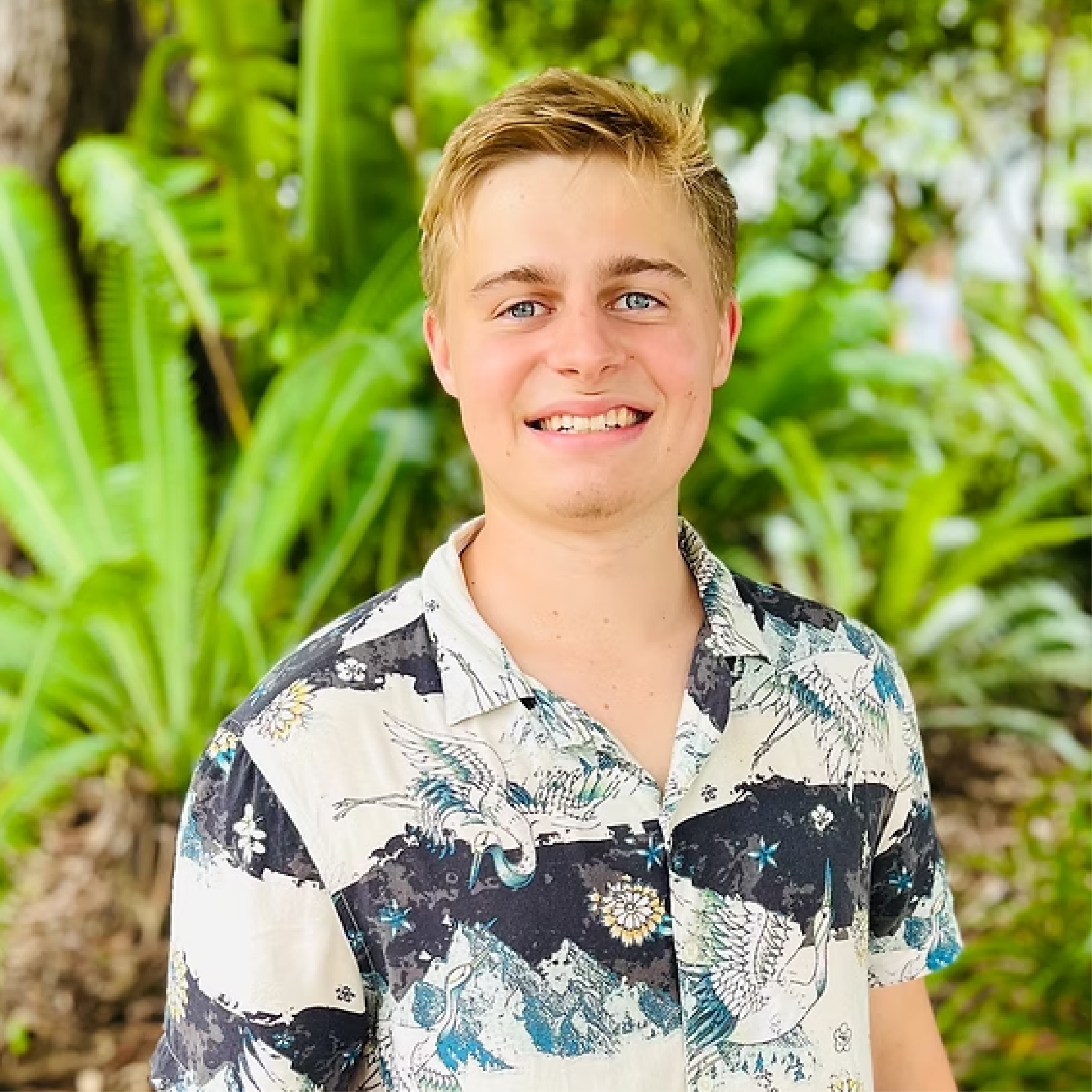 A young man smiling outdoors, wearing a Hawaiian shirt, with lush green tropical plants in the background.
