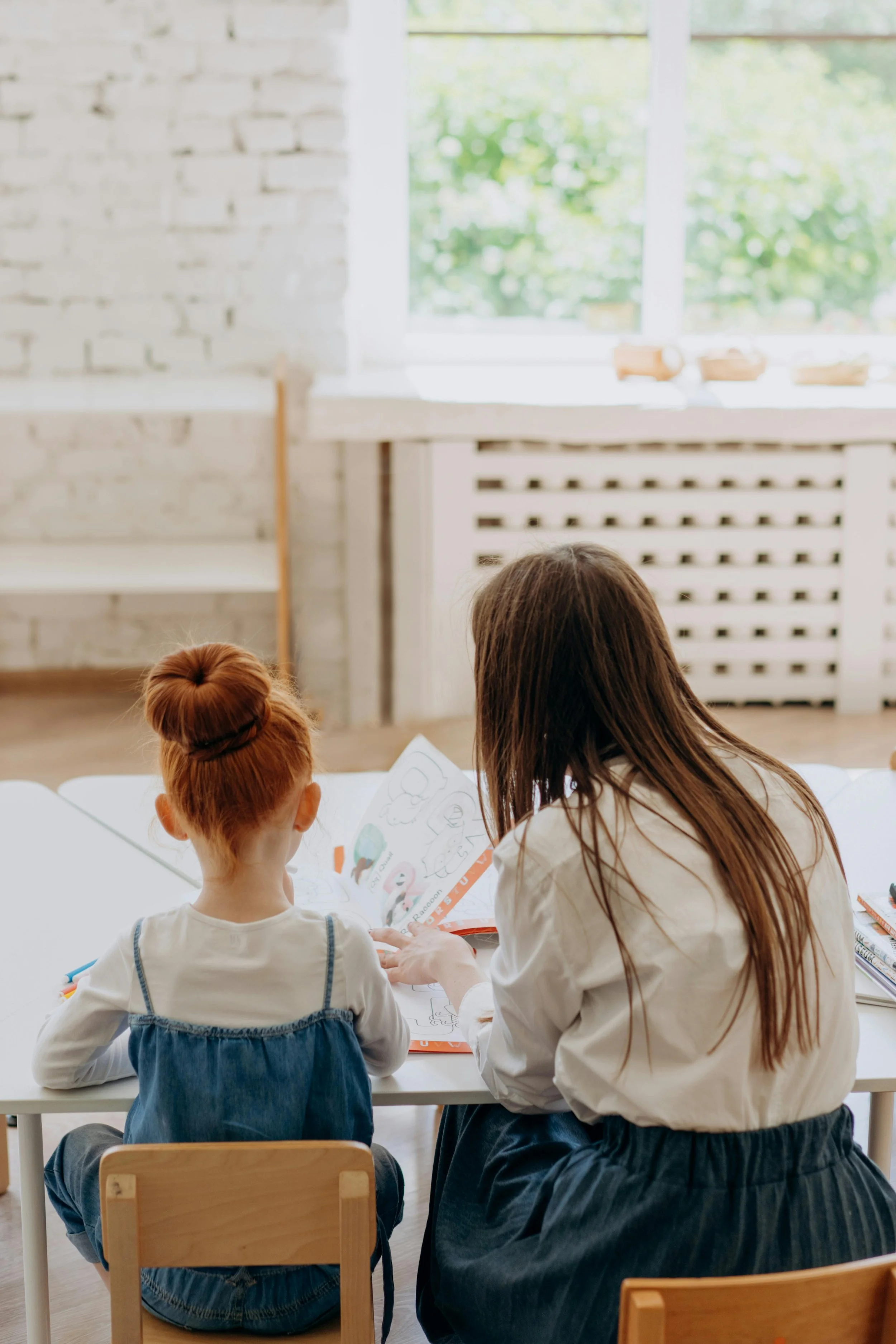 Adult and young girl sitting at a white table, coloring or reading a book, in a bright room with a large window and white brick wall.