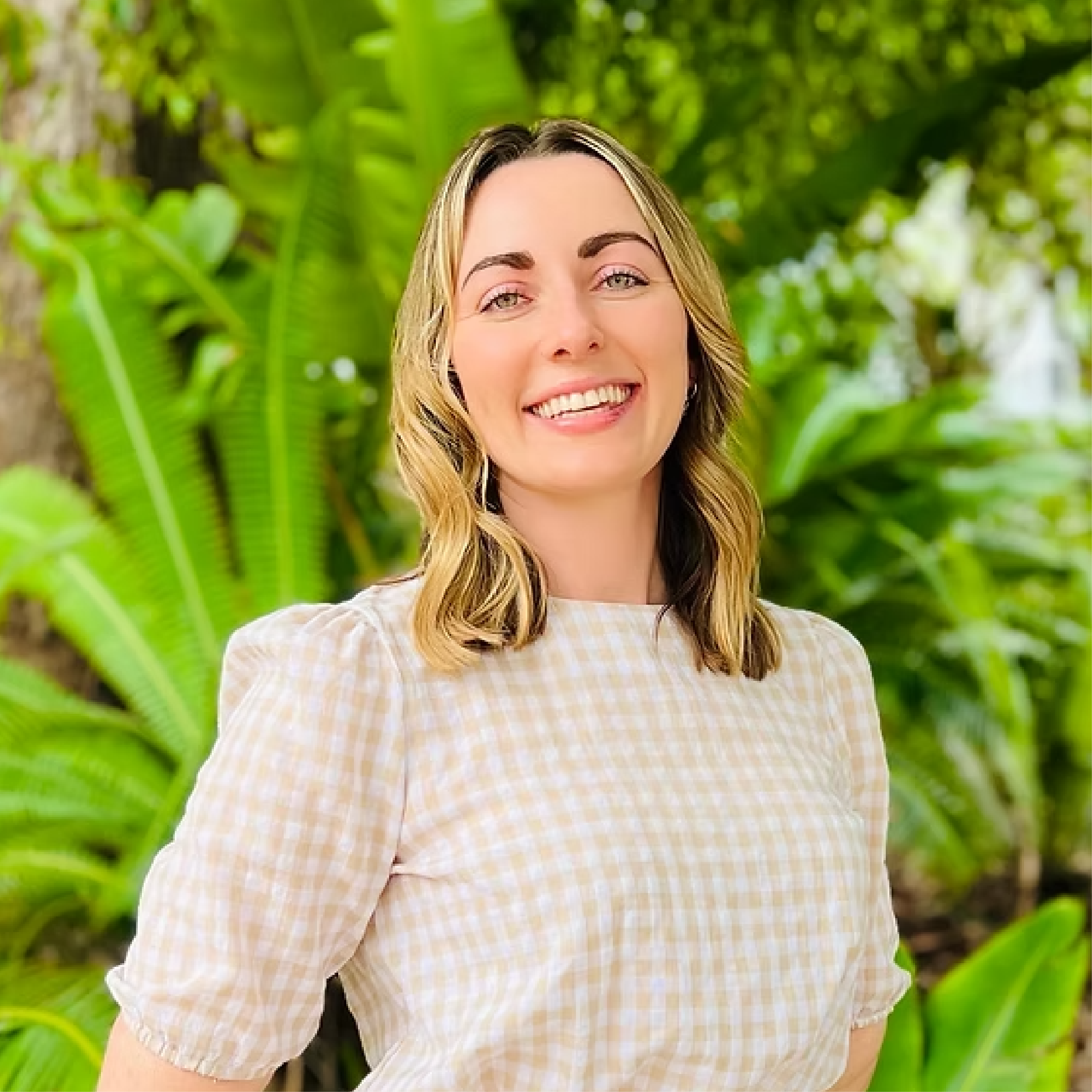 A woman with shoulder-length blonde hair, smiling, standing outdoors in front of lush green tropical plants.