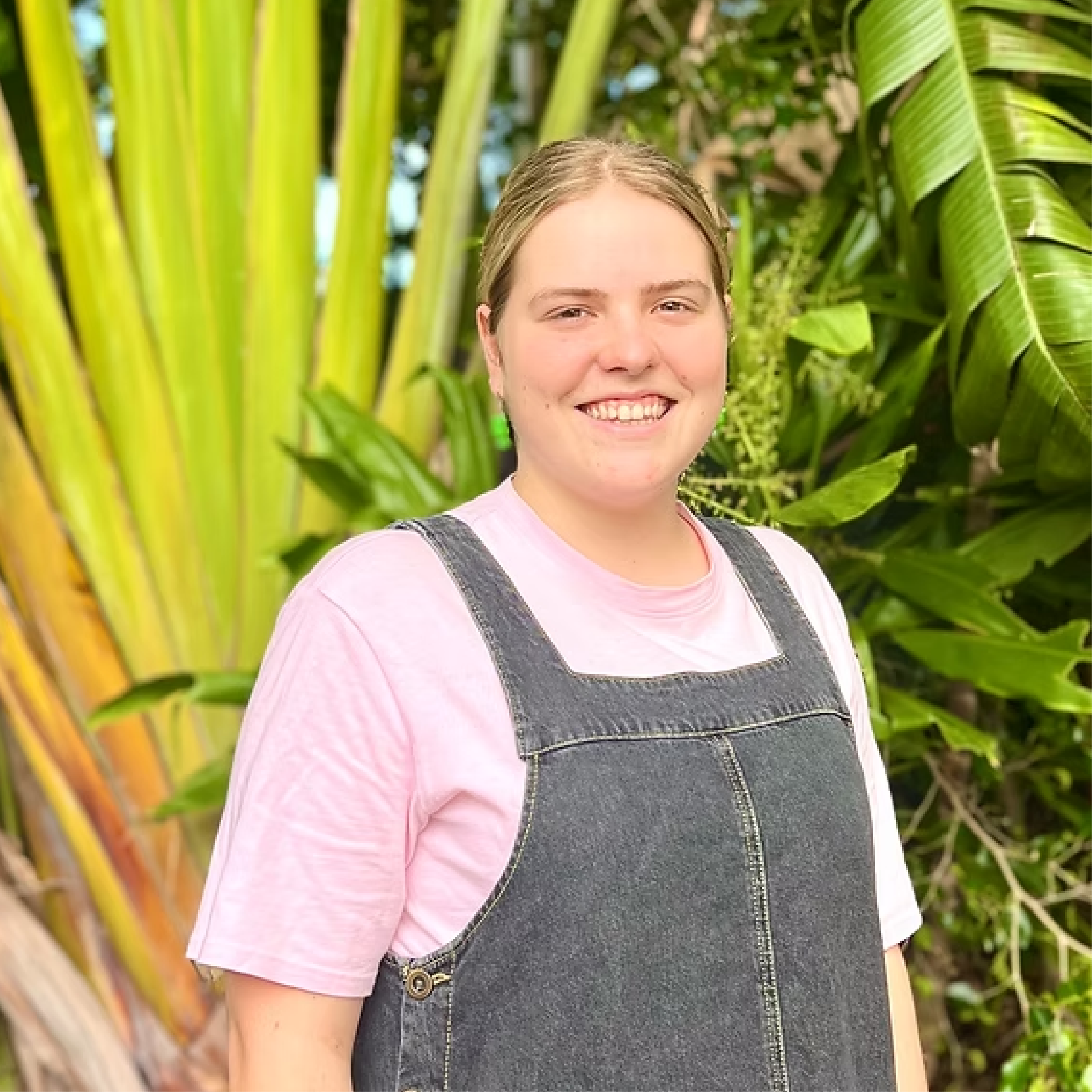 Young woman smiling outdoors in front of tropical green plants, wearing a pink shirt and dark denim overalls.