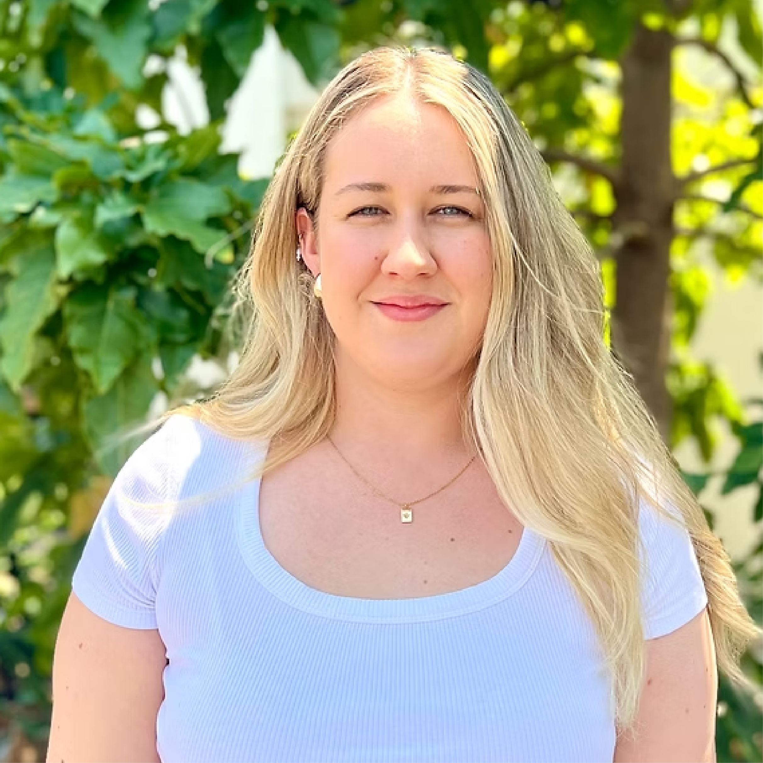 A young woman with long blonde hair, wearing a white shirt and a gold necklace, standing outdoors in front of green foliage and a tree, smiling gently.