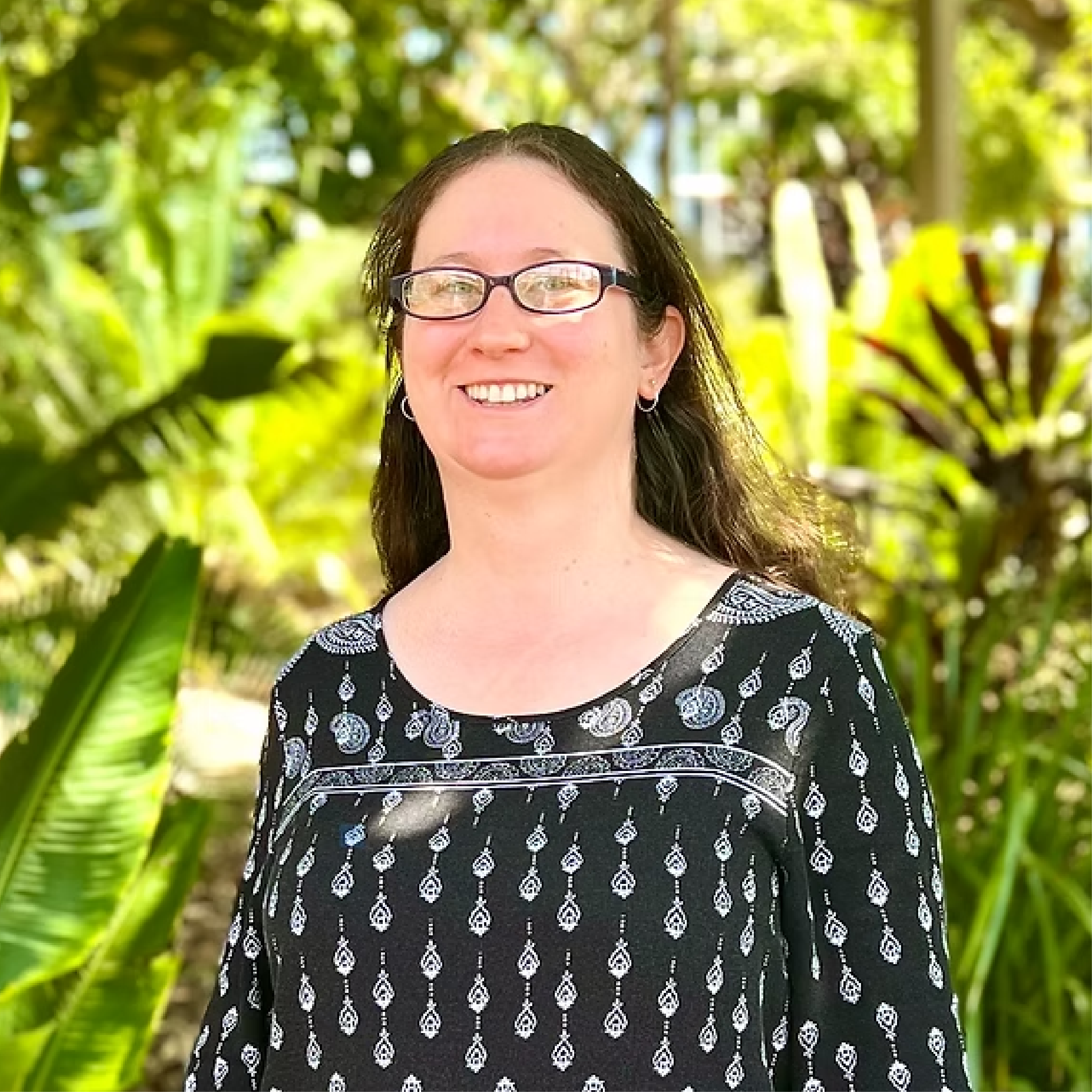 Woman with glasses smiling outdoors in a garden or park with green foliage in the background.