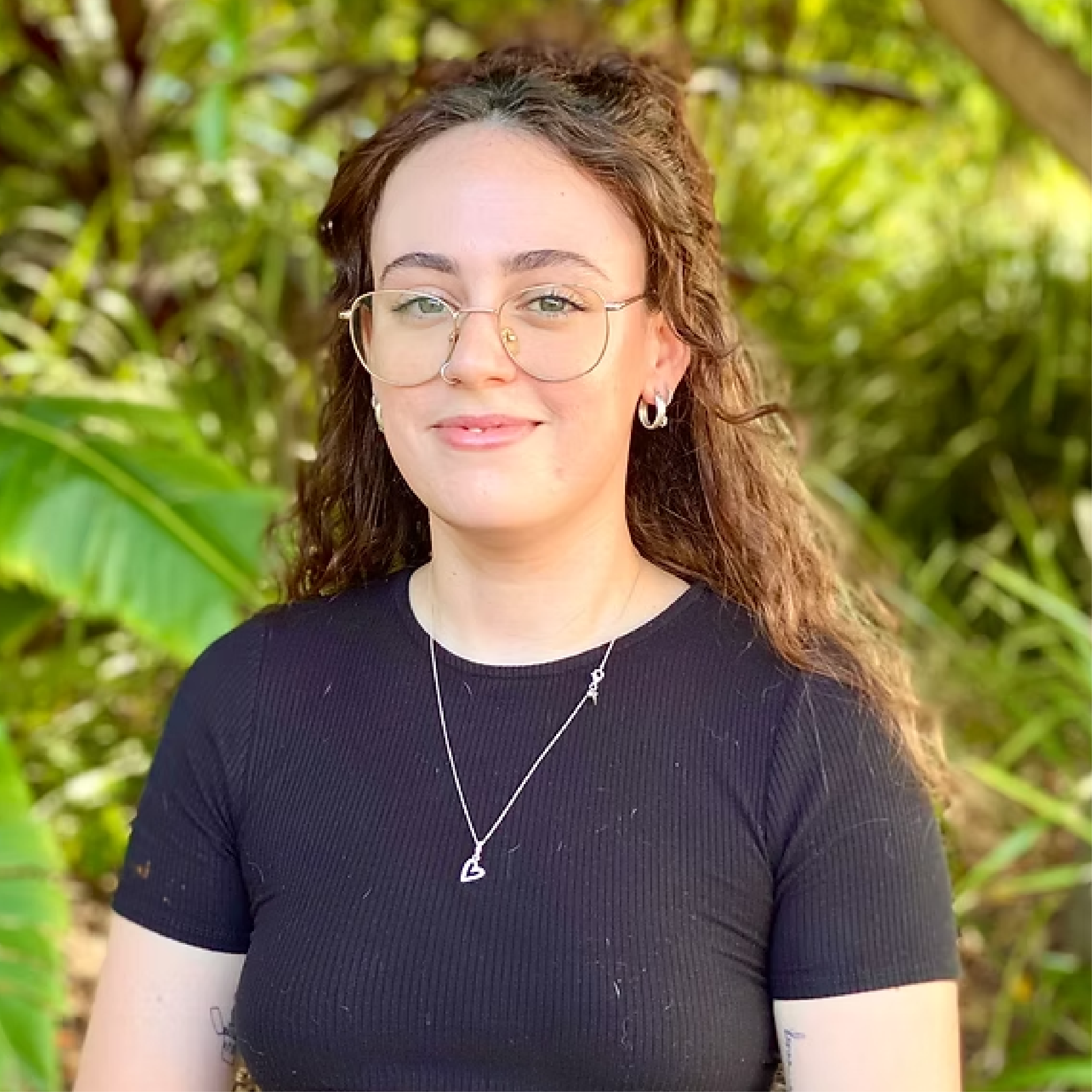 A young woman with curly brown hair, glasses, and multiple piercings wearing a black shirt and silver jewelry, standing outdoors in a lush green environment.
