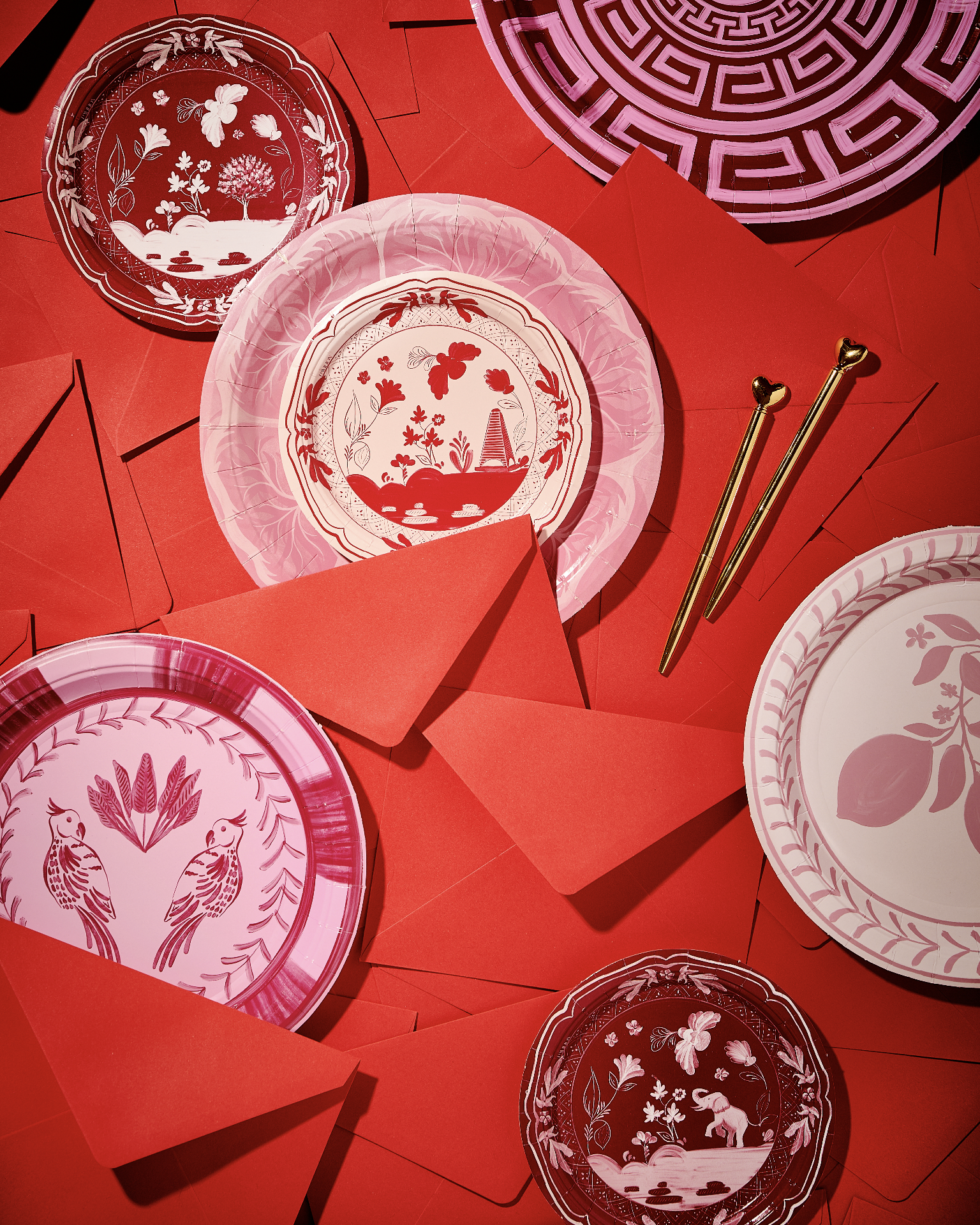 Assorted decorative plates with red and pink patterns, and red envelopes, on a red surface with two golden pens.