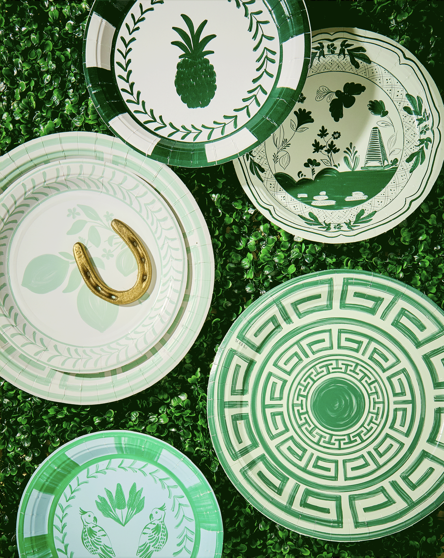 Multiple decorative plates with green and white patterns, some featuring pineapples, parrots, floral designs, and Greek key motifs, arranged on a green textured surface.