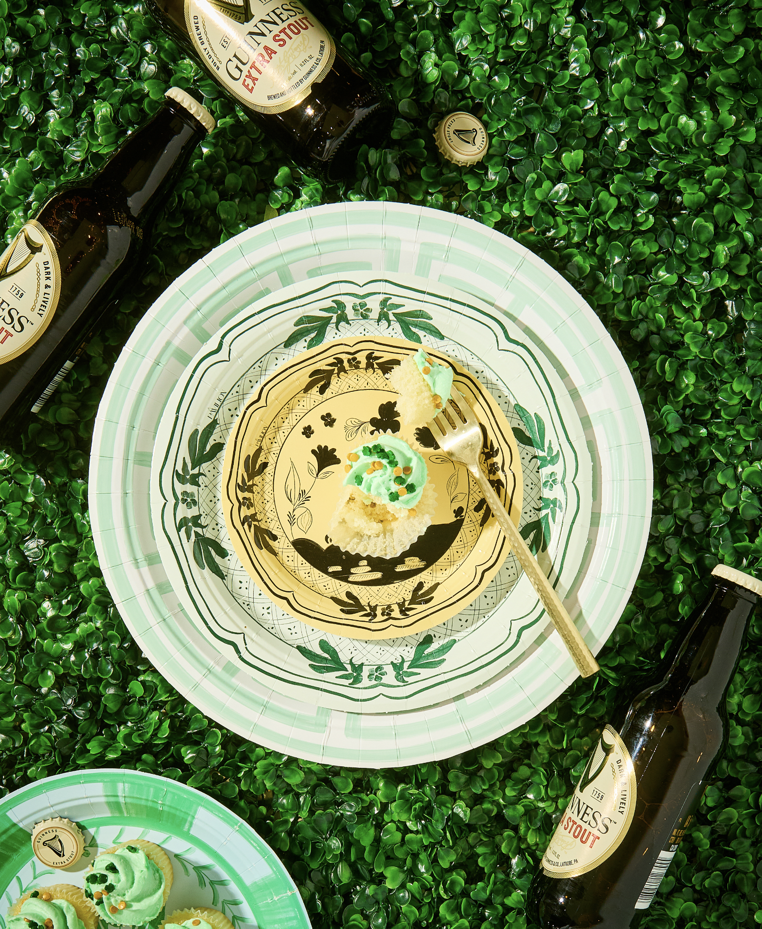 Cupcakes with green frosting and sprinkles on decorative plates surrounded by Guinness bottles on artificial grass.