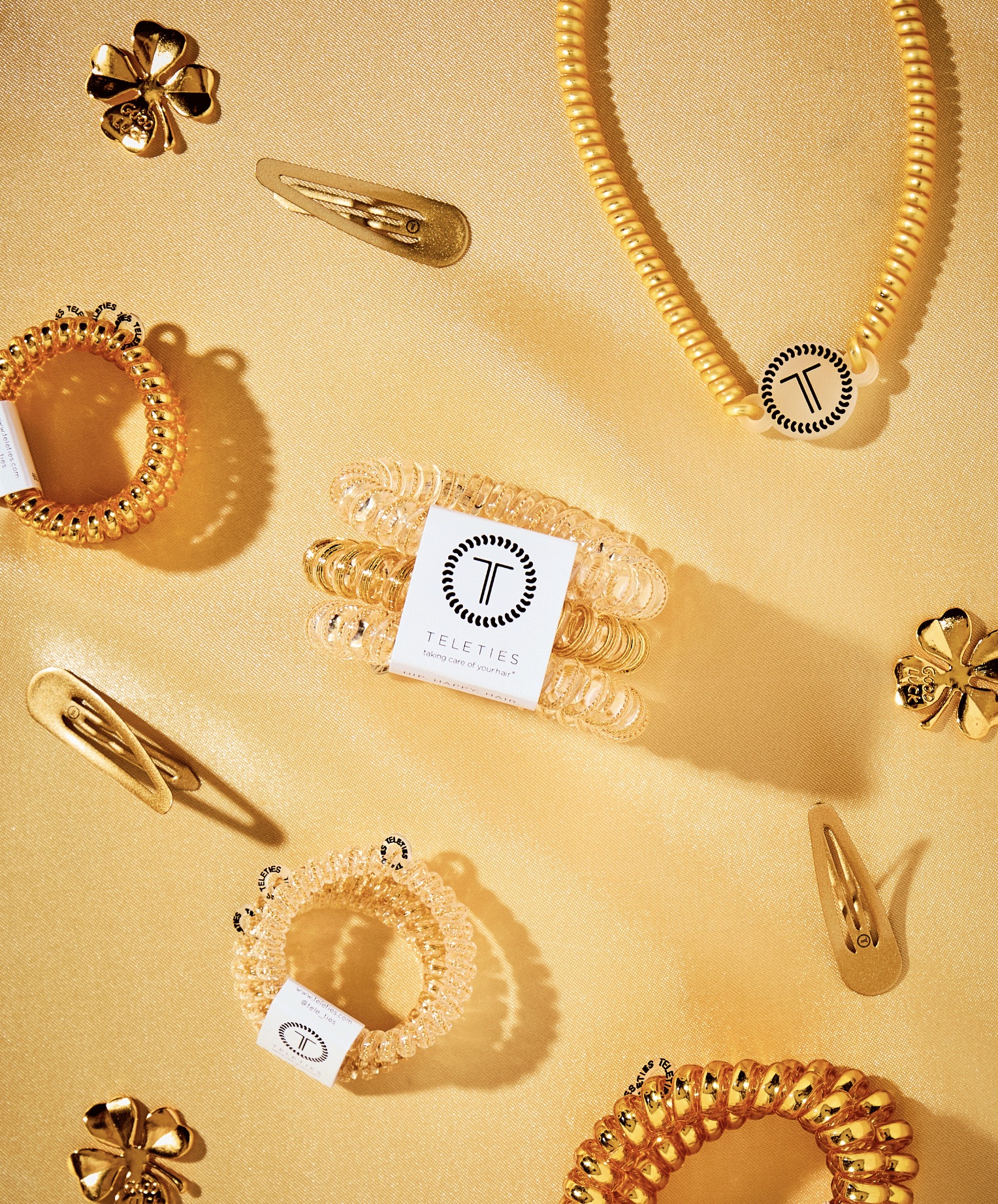 Collection of gold-colored hair accessories and jewelry, including hair clips and scrunchies, arranged on a gold fabric background.