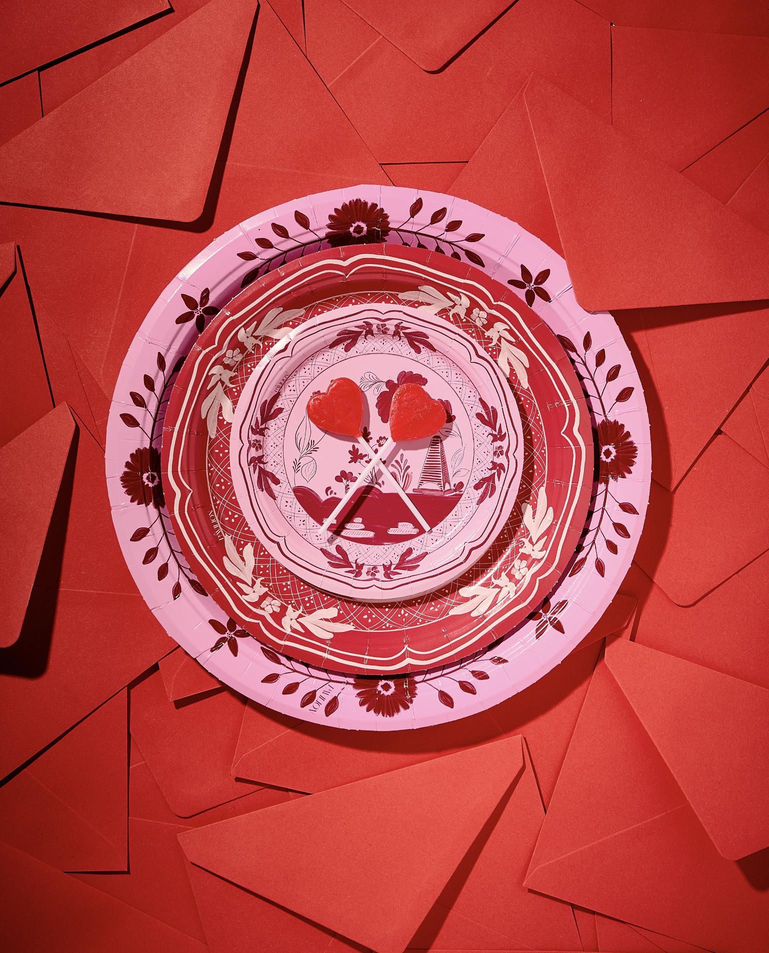 Two red heart-shaped lollipops on sticks resembling a couple kissing, placed on a decorative red and pink paper plate with floral and nature patterns, on a background of scattered red envelopes.