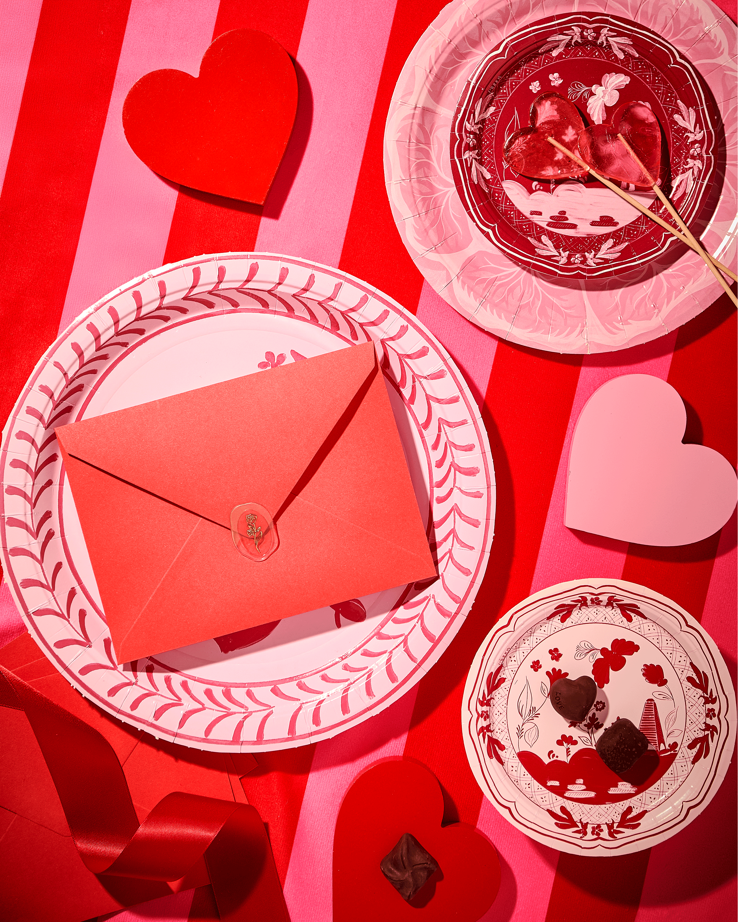 Valentine's Day themed table setting with pink and red hearts, a red envelope on a decorative plate, heart-shaped lollipops, chocolates, and red and pink striped background.
