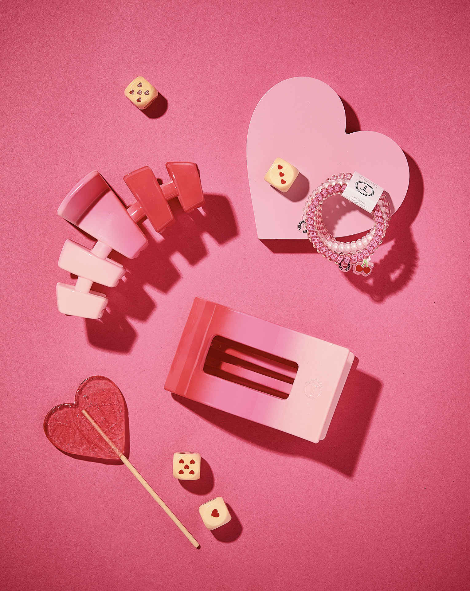Pink background with pink and red hair clips, a pink and white beaded bracelet with cherry charms, a pink heart-shaped paper with a label, a pink stamp, a heart-shaped lollipop, and two small dice with red hearts.