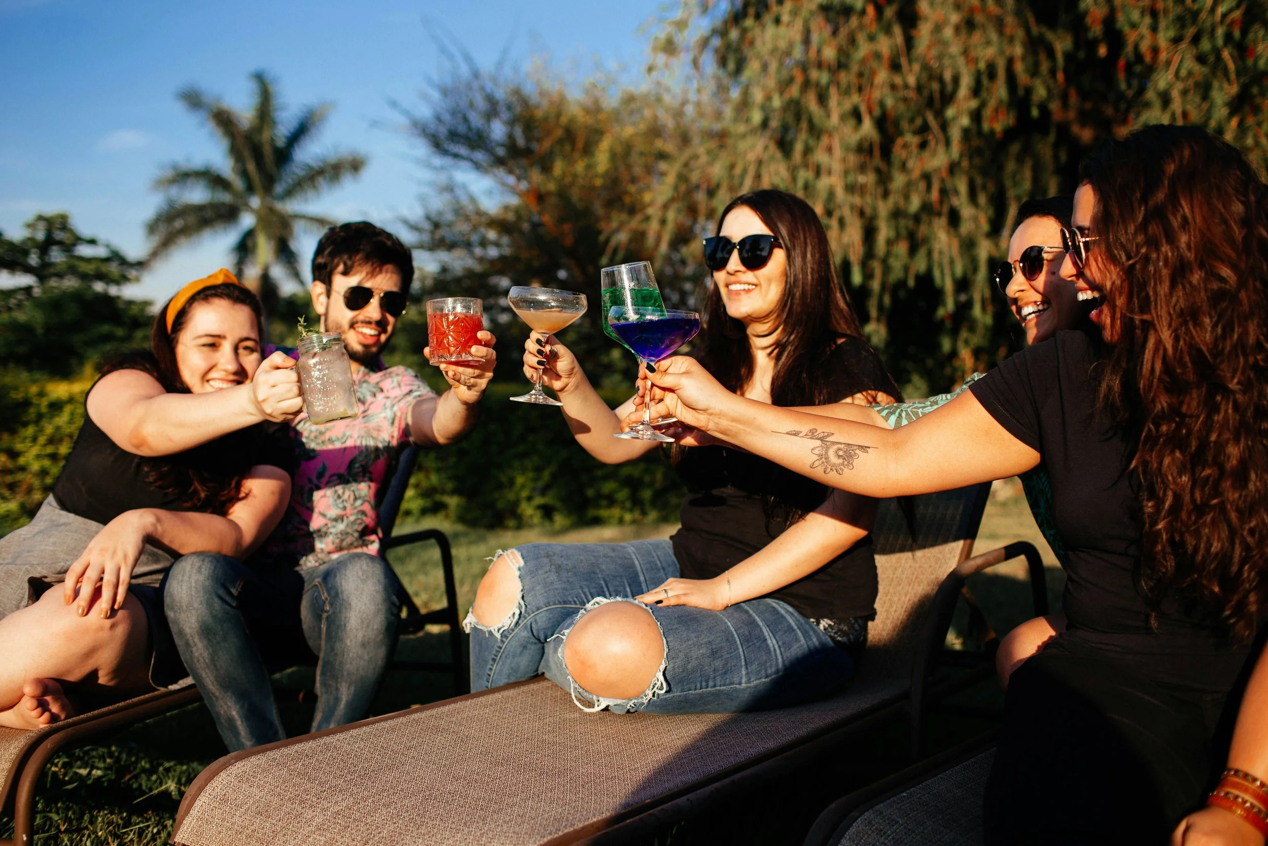 Group of friends enjoying cocktails outdoors, sitting on lounge chairs with trees and a clear sky in the background, celebrating together.