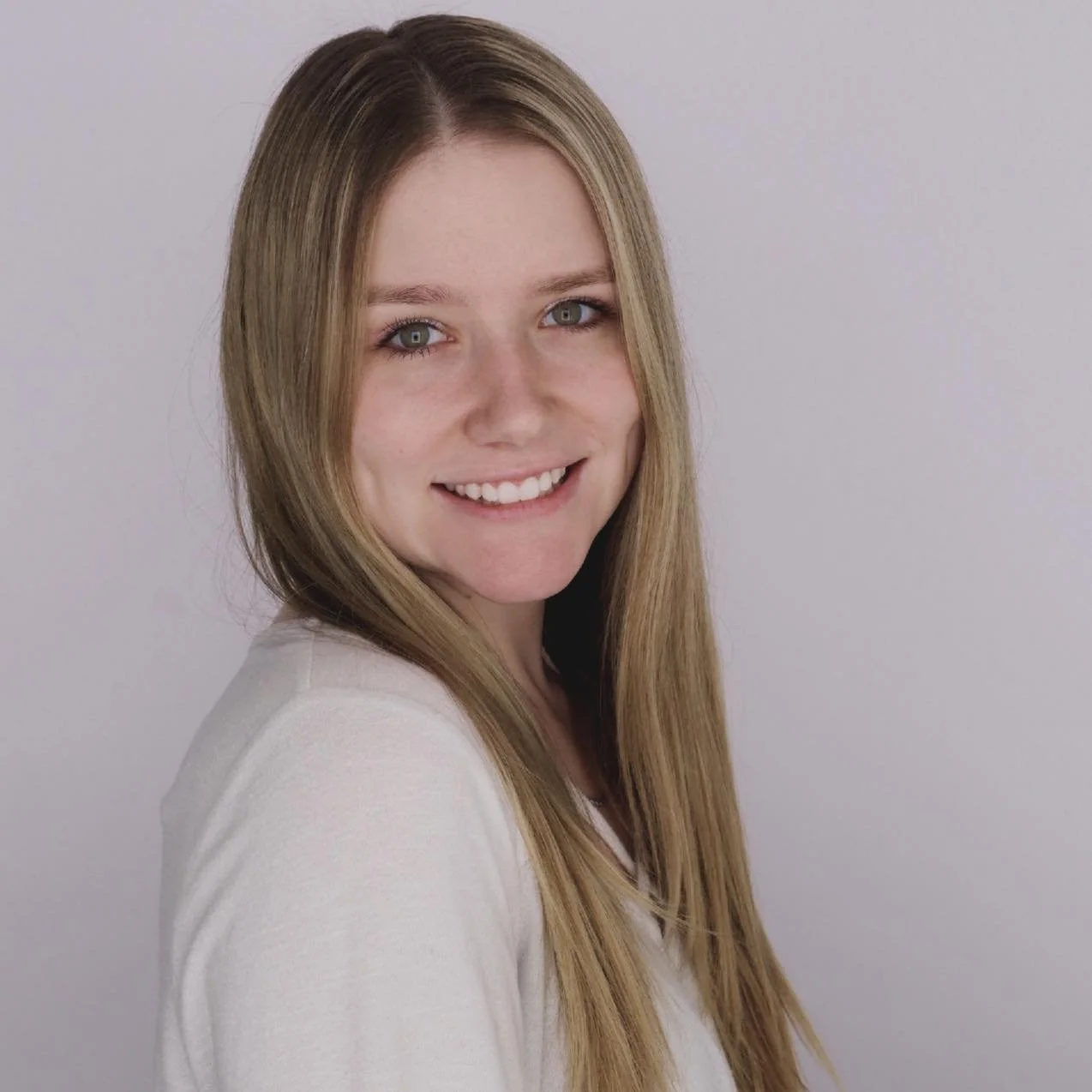 Portrait of a young woman with long blonde hair smiling, wearing a white top, against a plain light gray background.