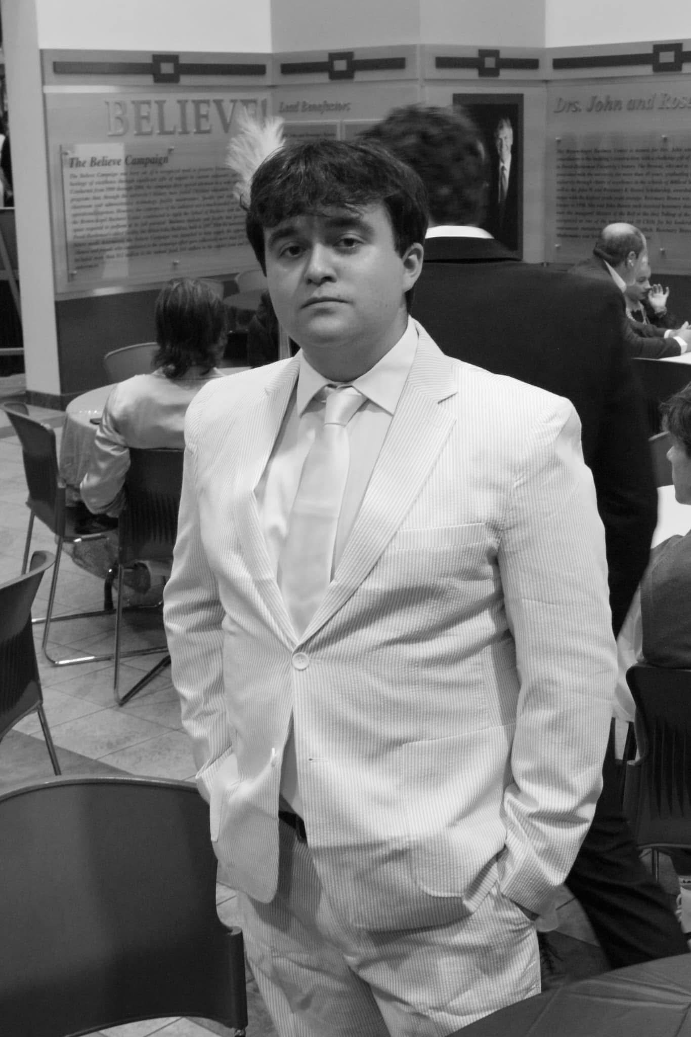 A young man in a light-colored suit and tie standing in an indoor event space, with several people seated behind him, including a woman with short hair and a man with glasses, and displayed posters and photos on the wall.