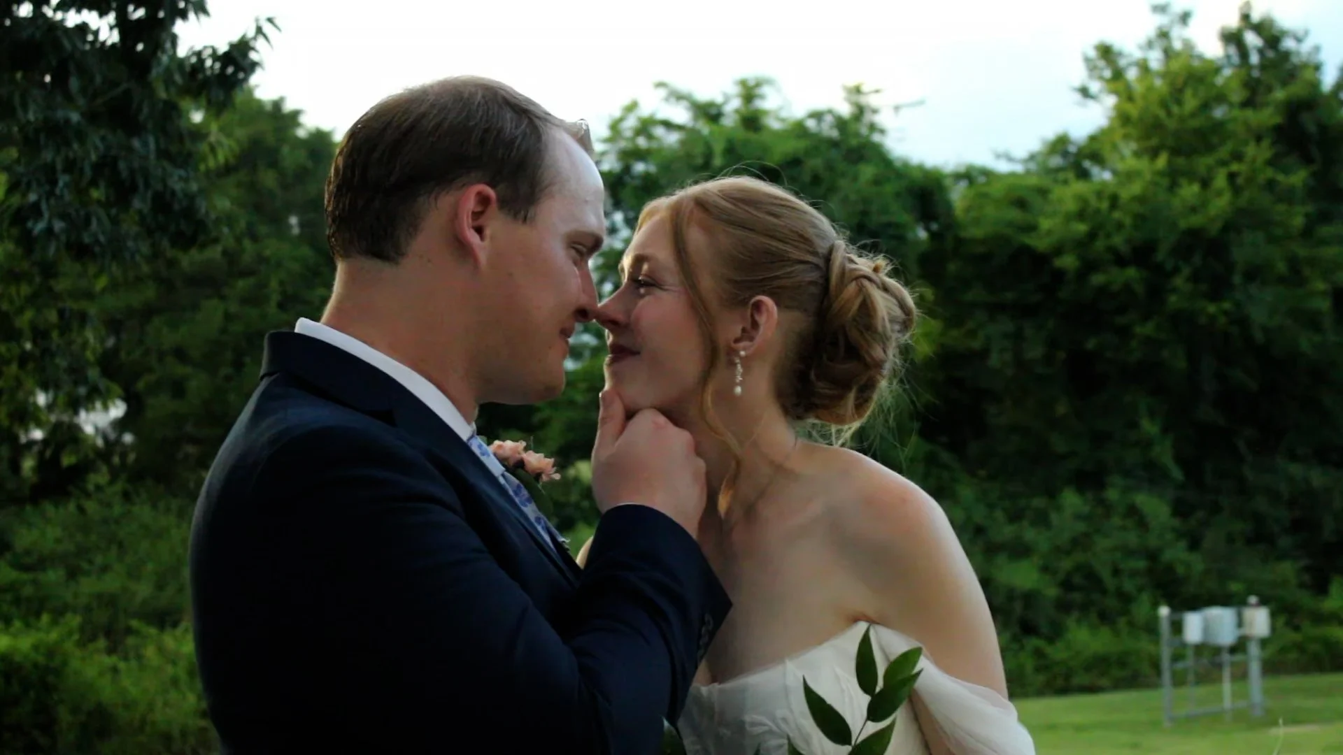 A man and woman on their wedding day, standing close together outdoors, smiling and touching noses, surrounded by green trees.