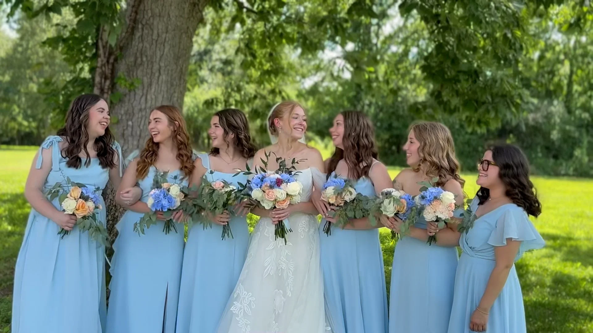 A bridal party of eight women in light blue dresses, standing outdoors under a large tree, holding bouquets, and smiling at each other.