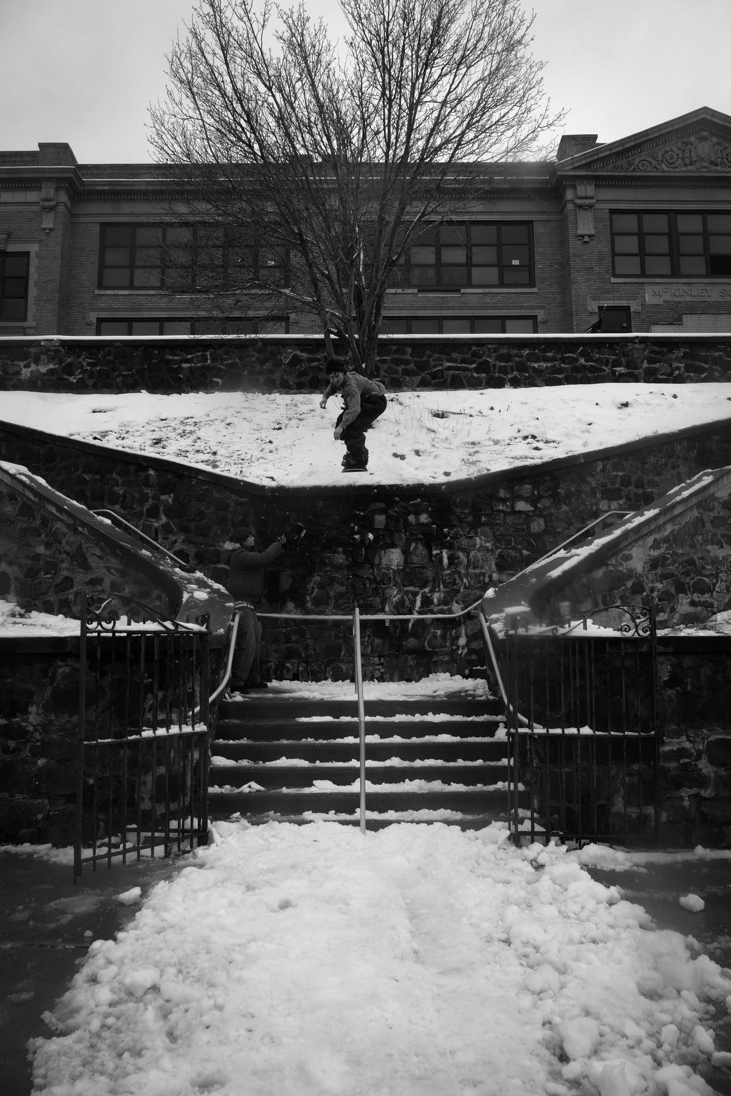 Child wearing winter clothing is sliding down a snowy slope while another person stands at the bottom. Snow covers the stairs and ground, with a large leafless tree and a brick building in the background.