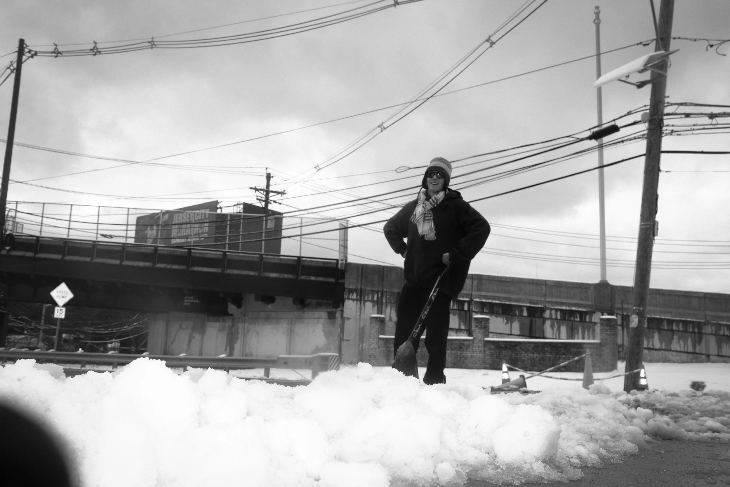 Person standing in snow with a shovel, wearing a hat and sunglasses, near a utility pole and power lines in an urban area with an overpass and cloudy sky.