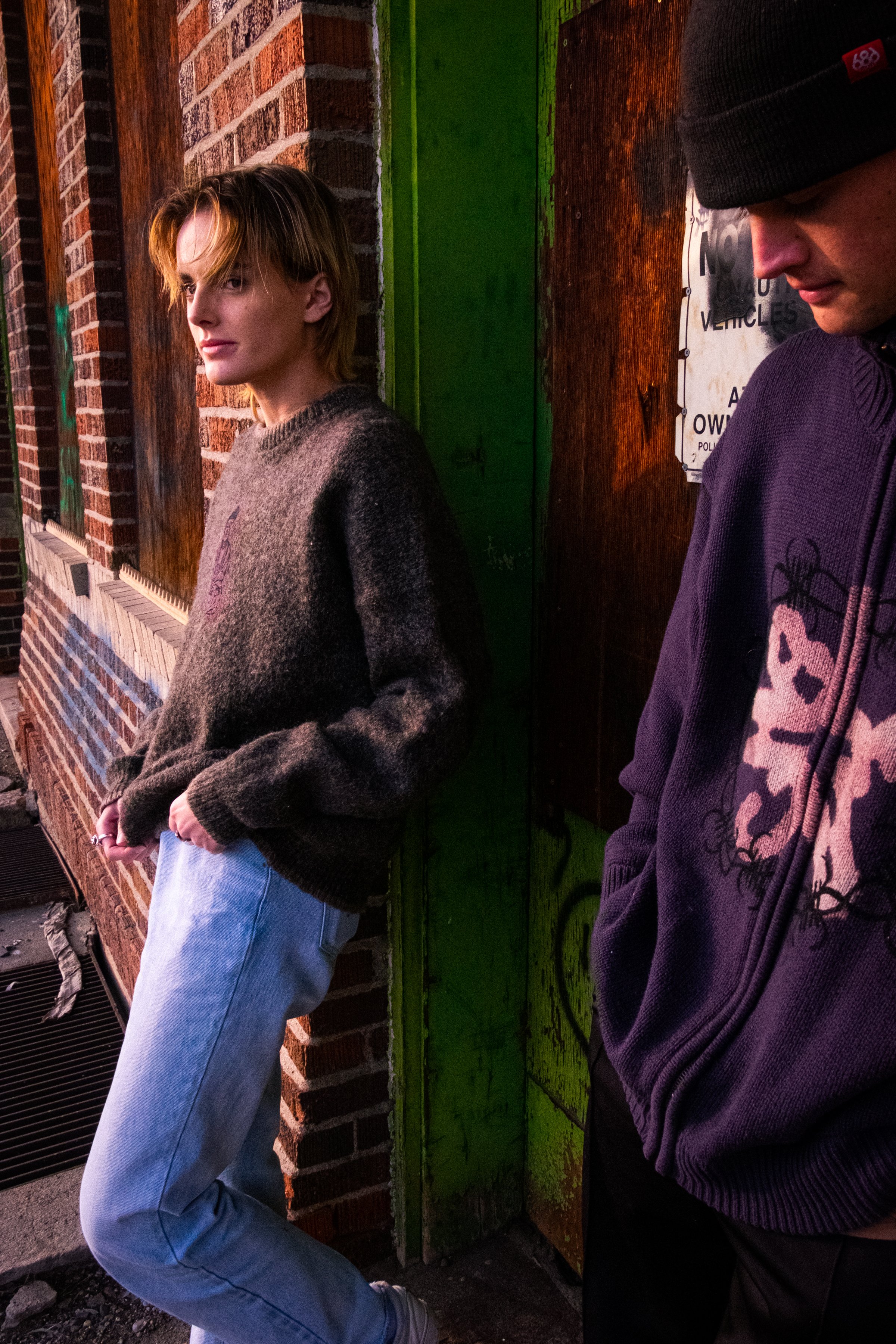 Two young men standing against a brick wall illuminated by streetlights at night, one looking into the camera and the other looking down.