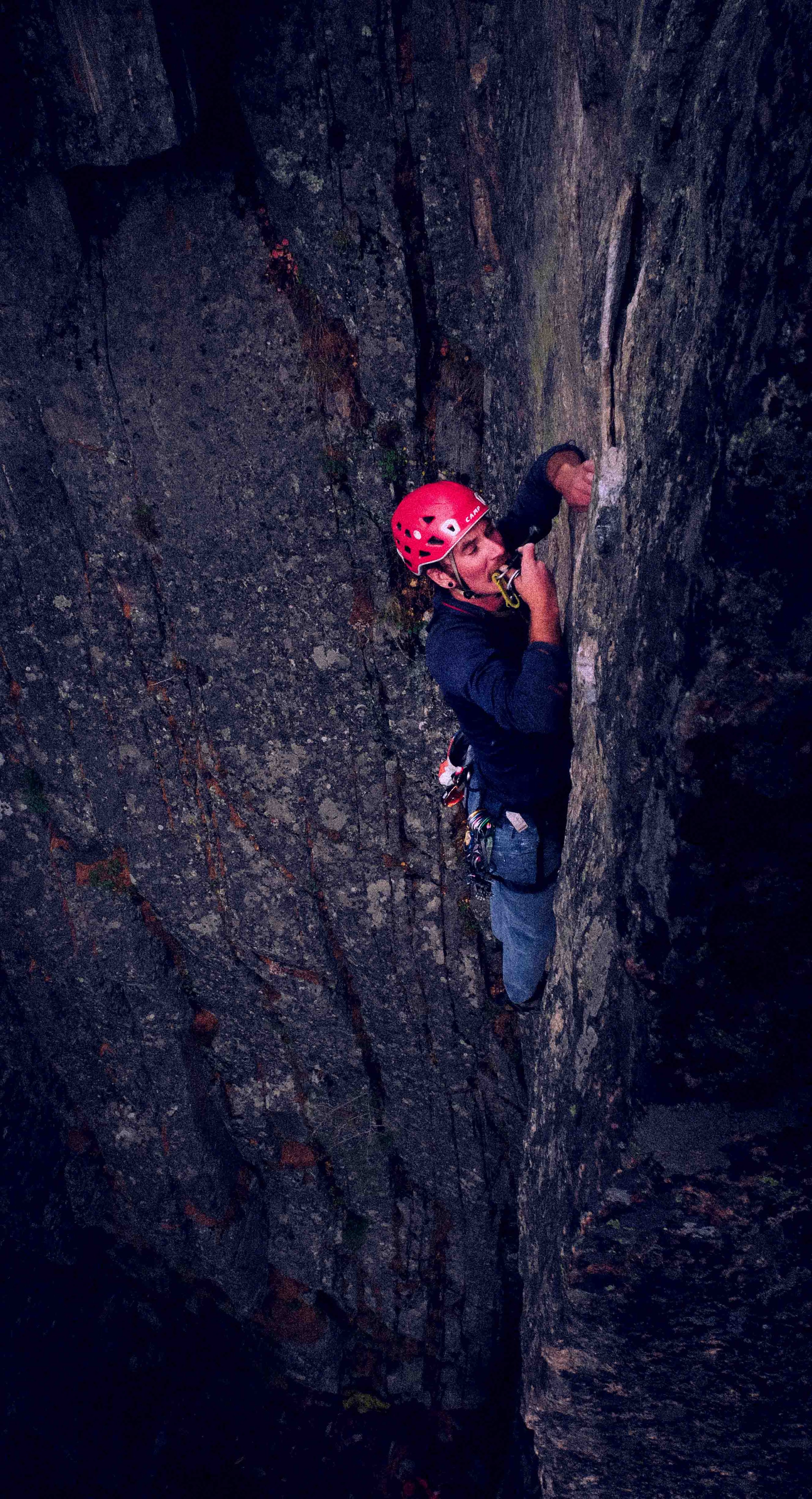 A man wearing a red climbing helmet and harness is rock climbing on a vertical cliff face at dusk or dawn.