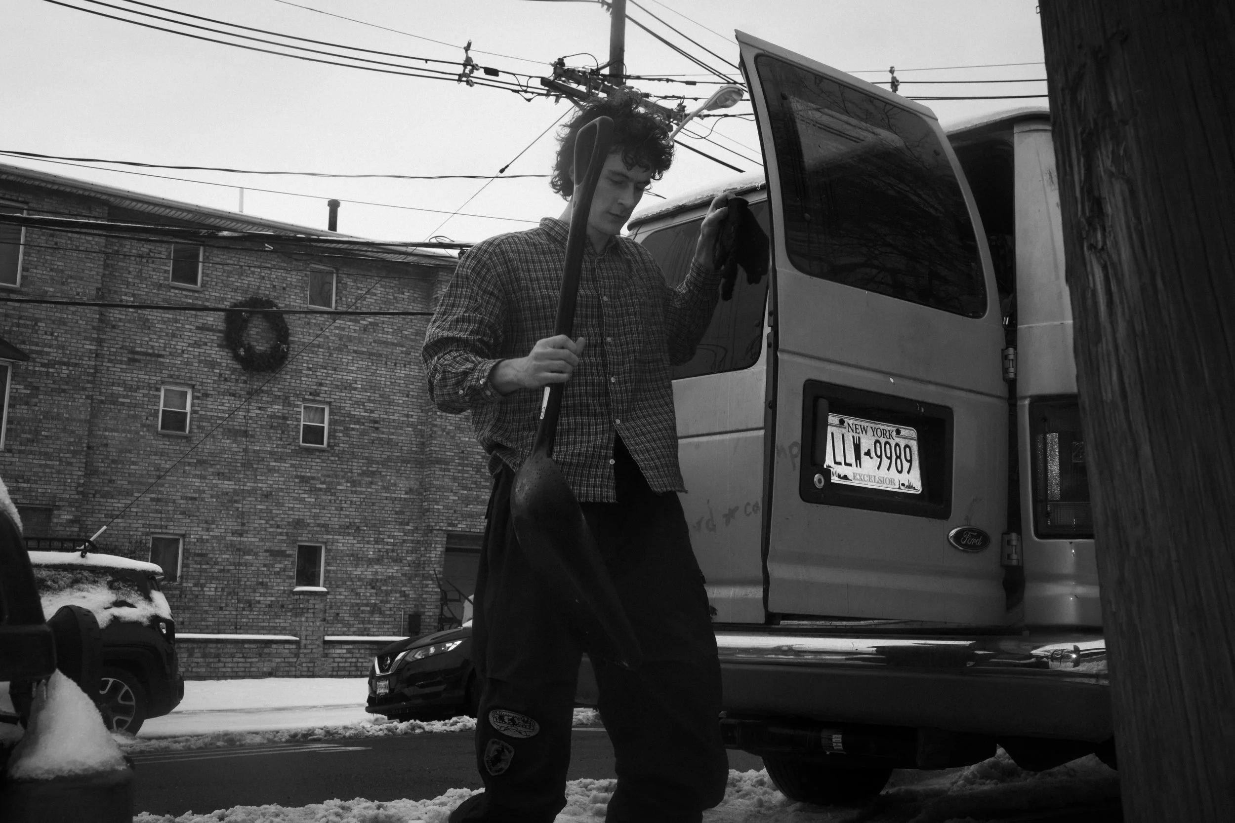 A young man with curly hair and a checkered shirt holding a snow shovel, walking on a snowy street near a van and parked cars, with a brick building in the background and electrical wires above.