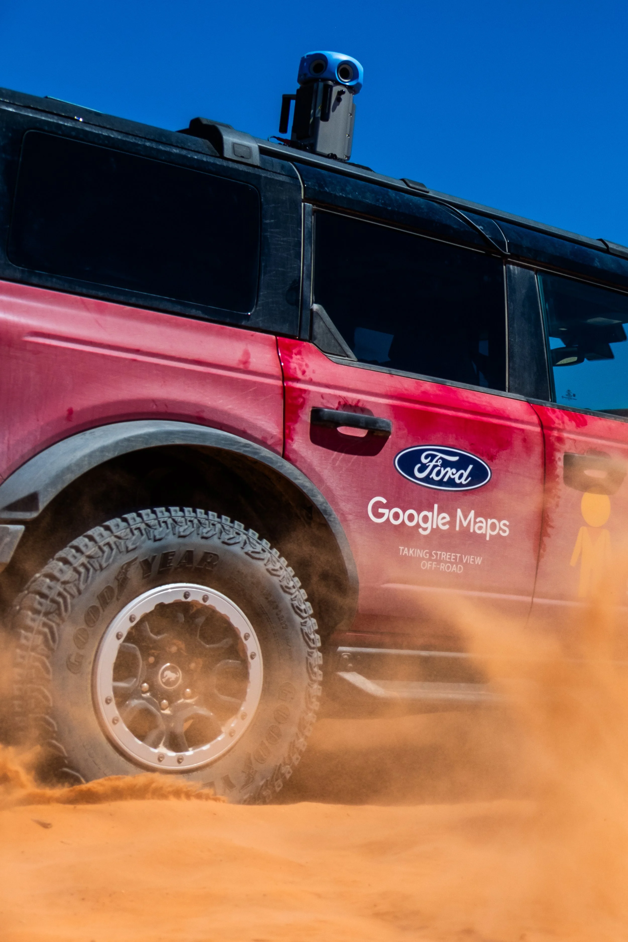 A red off-road vehicle with Google Maps branding on the side, driving through a sandy desert with dust and orange sand kicked up by the tires, against a clear blue sky.