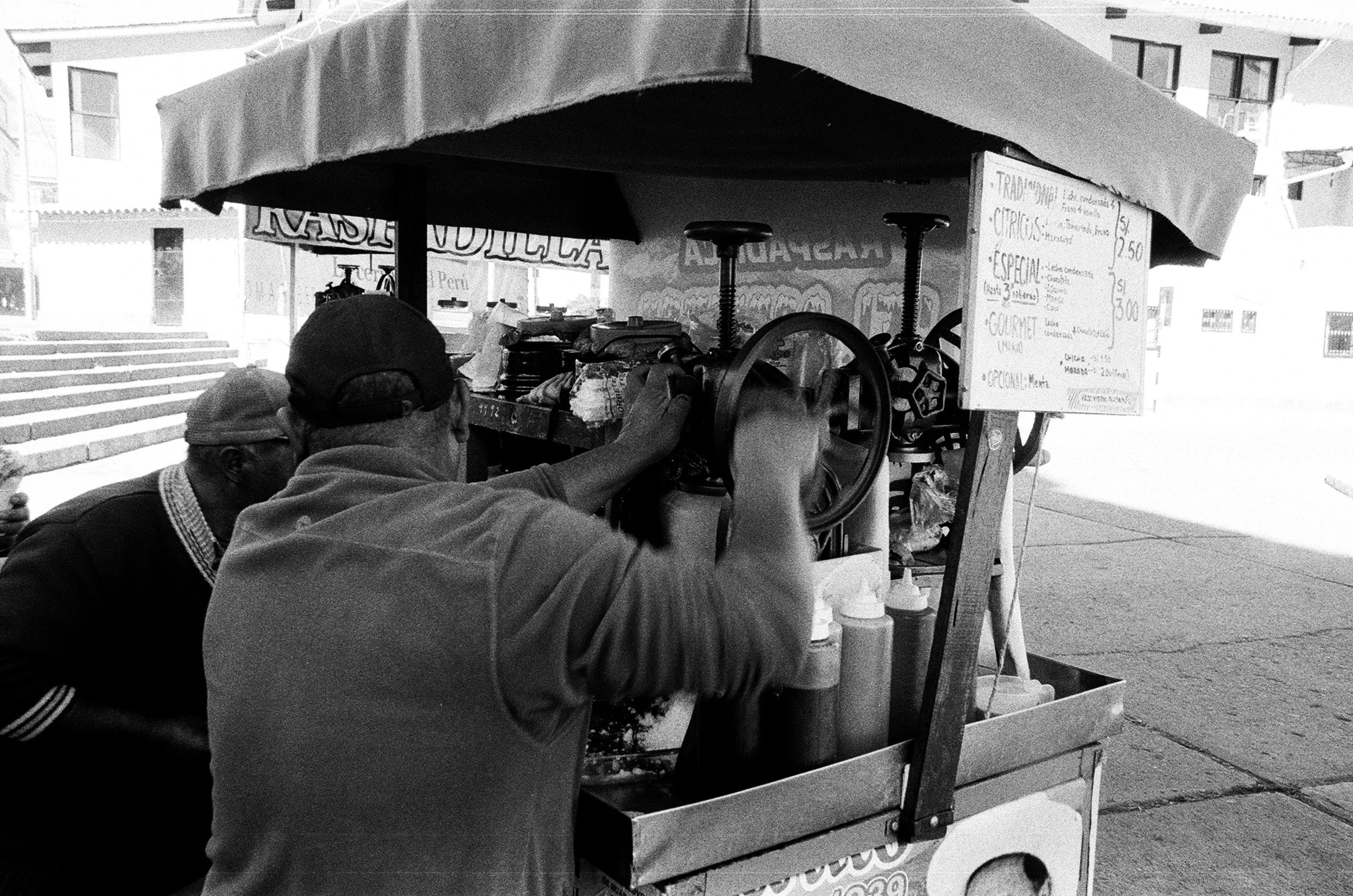 A street food cart serving drinks and snacks, with two men preparing orders, in an urban setting with stairs and buildings in the background.