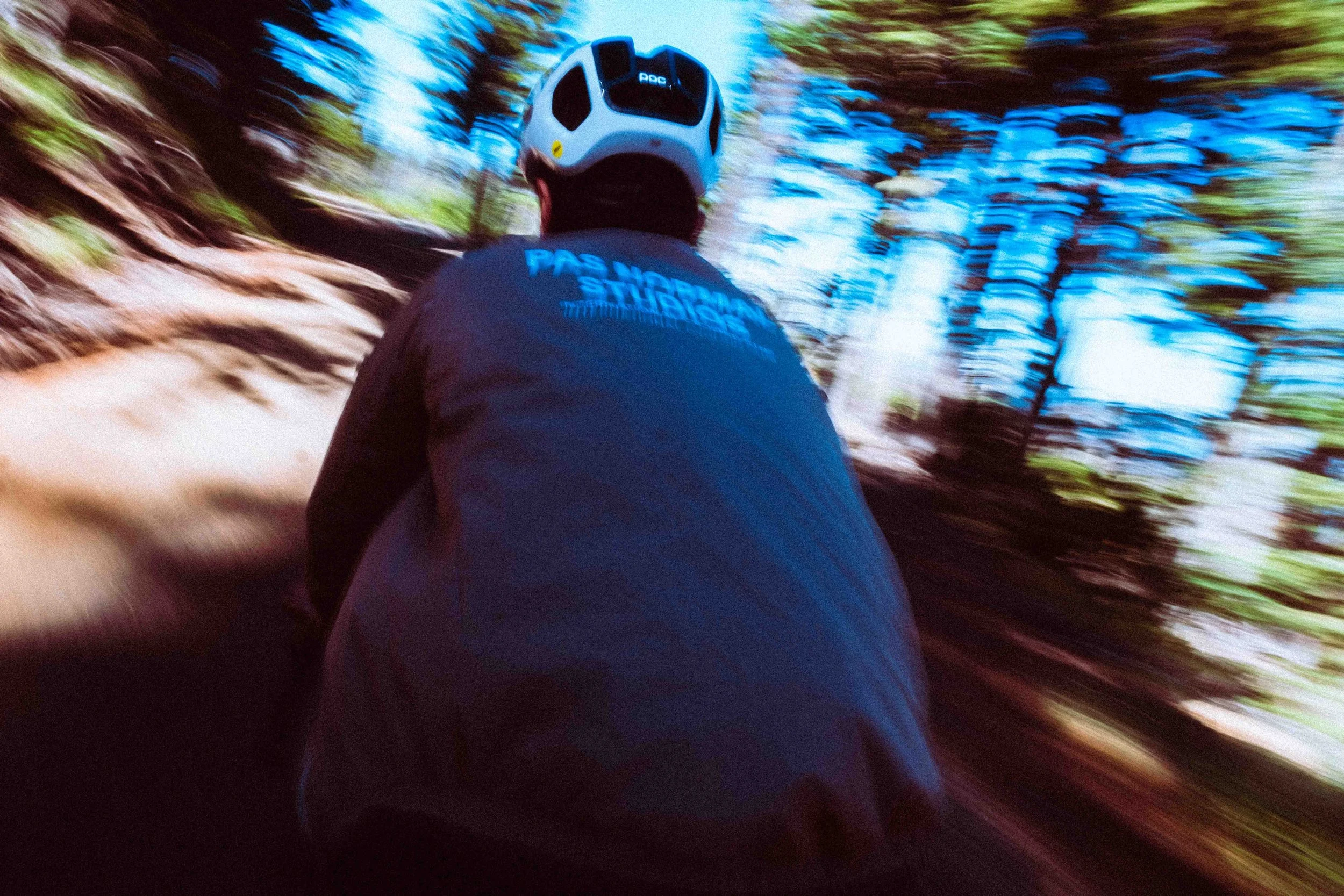 A person wearing a helmet and a dark jacket riding a mountain bike through a trail in a wooded area, with blurred trees around.
