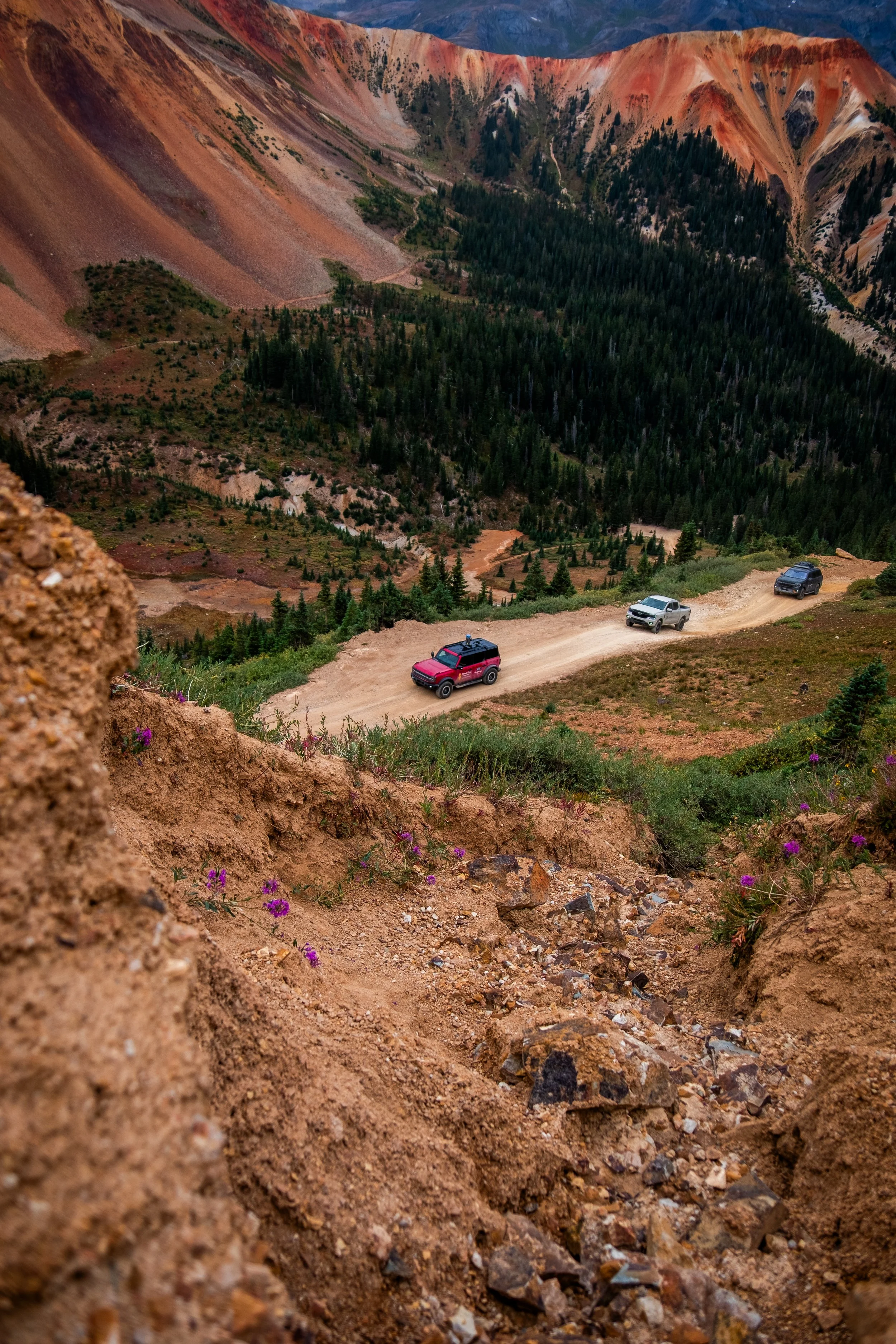 Three vehicles driving on a dirt mountain road with colorful, steep terrain and a forested valley in the background.