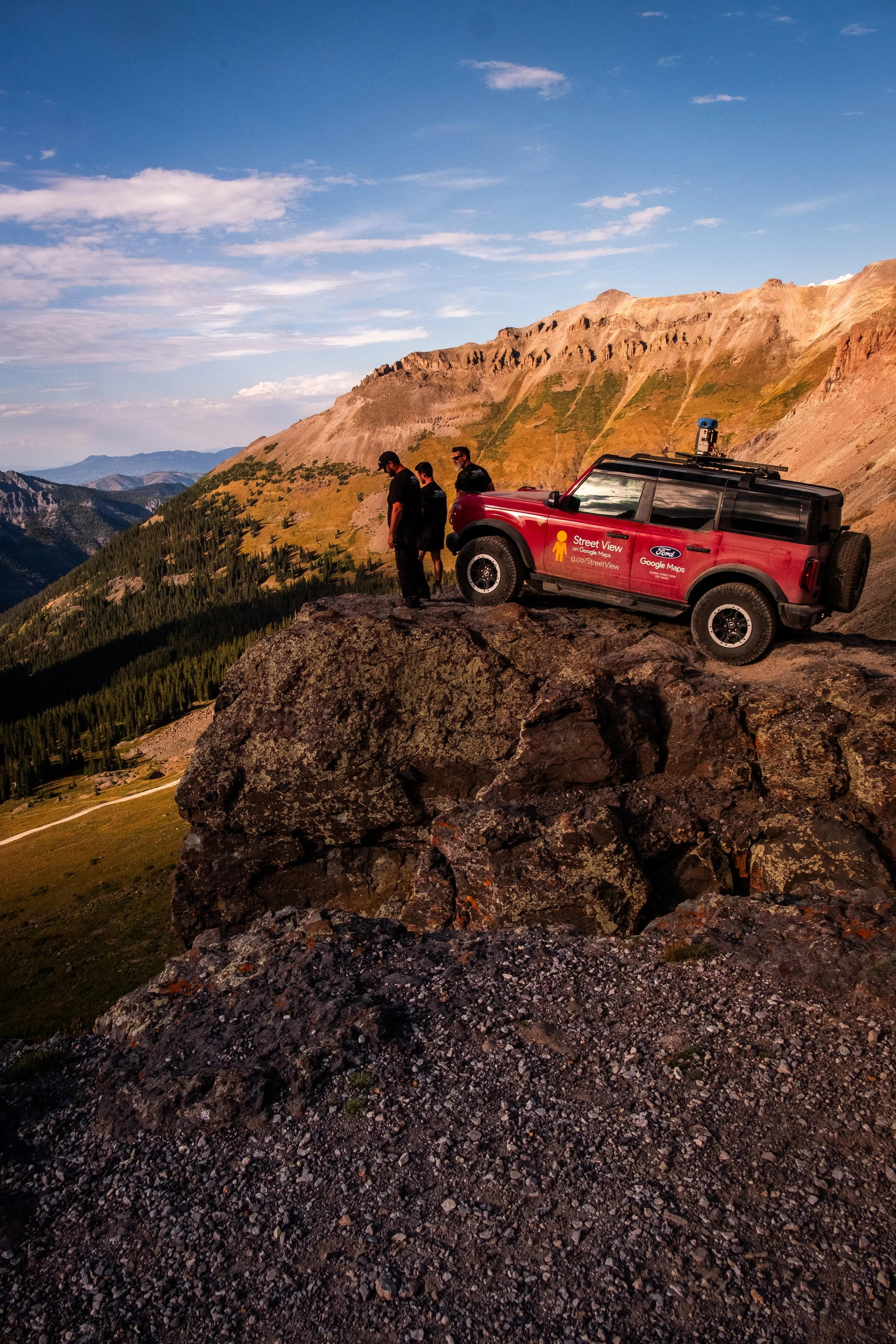 Three people standing on a large rock outcrop in a mountainous landscape during sunset, with a red off-road vehicle parked nearby.