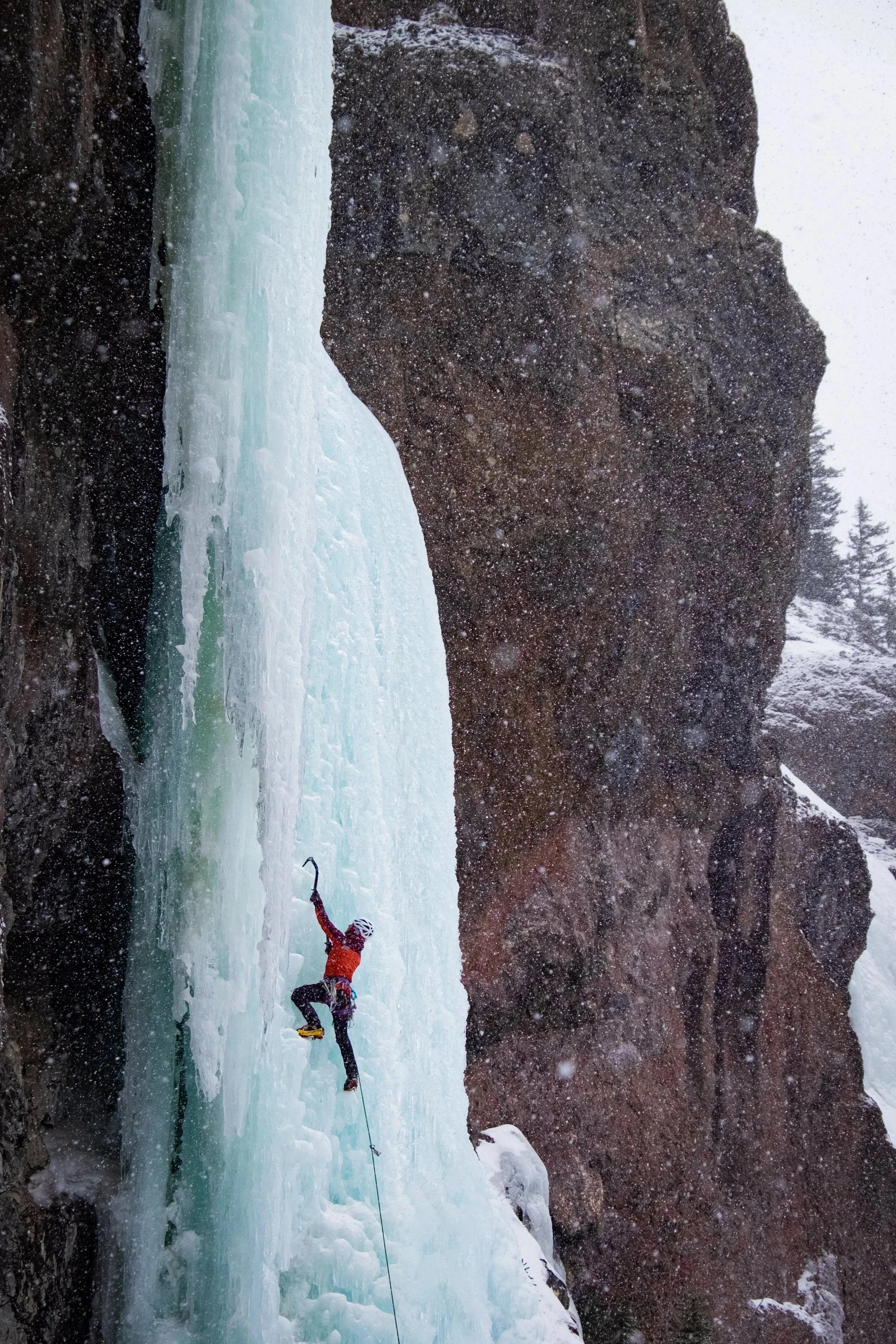 Ice climber wearing a red jacket and helmet ascending a frozen waterfall in a snowy, mountainous landscape.