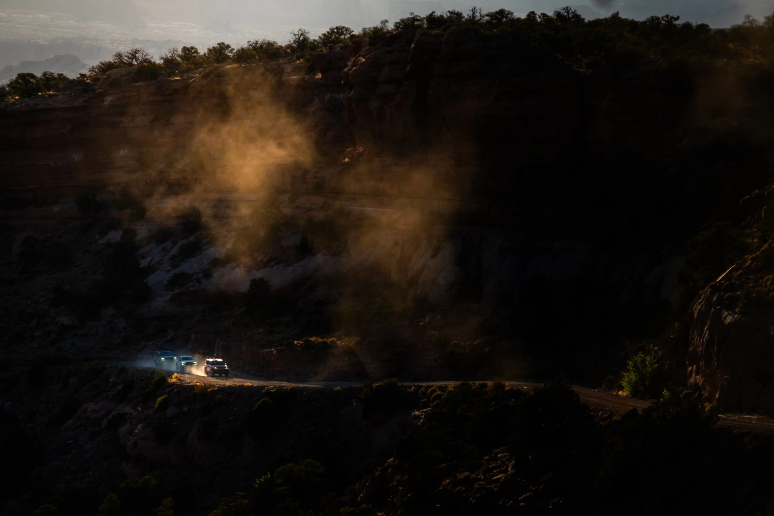 Four vehicles driving on a winding mountain road with dust clouds and rocky cliffs, illuminated by late afternoon or early evening sunlight.