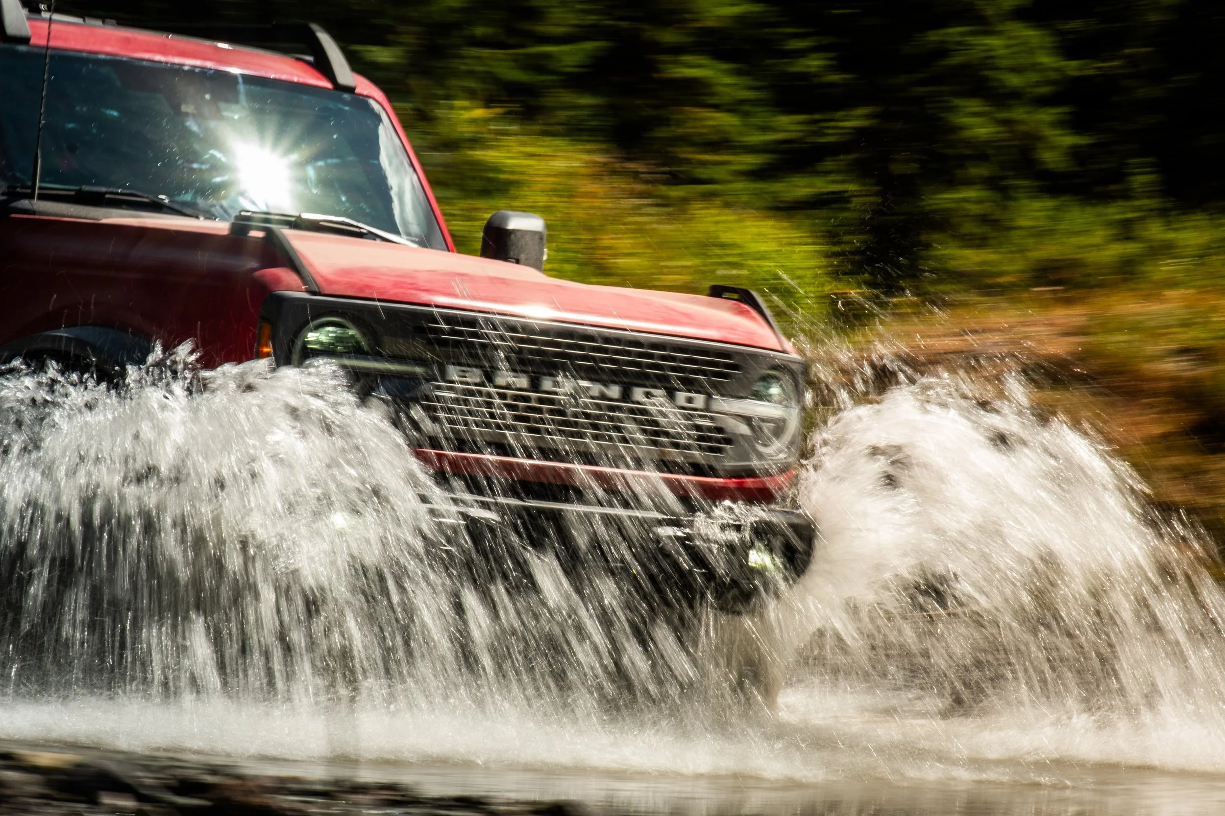 A red SUV driving through water, splashing water around with a blurred green background.