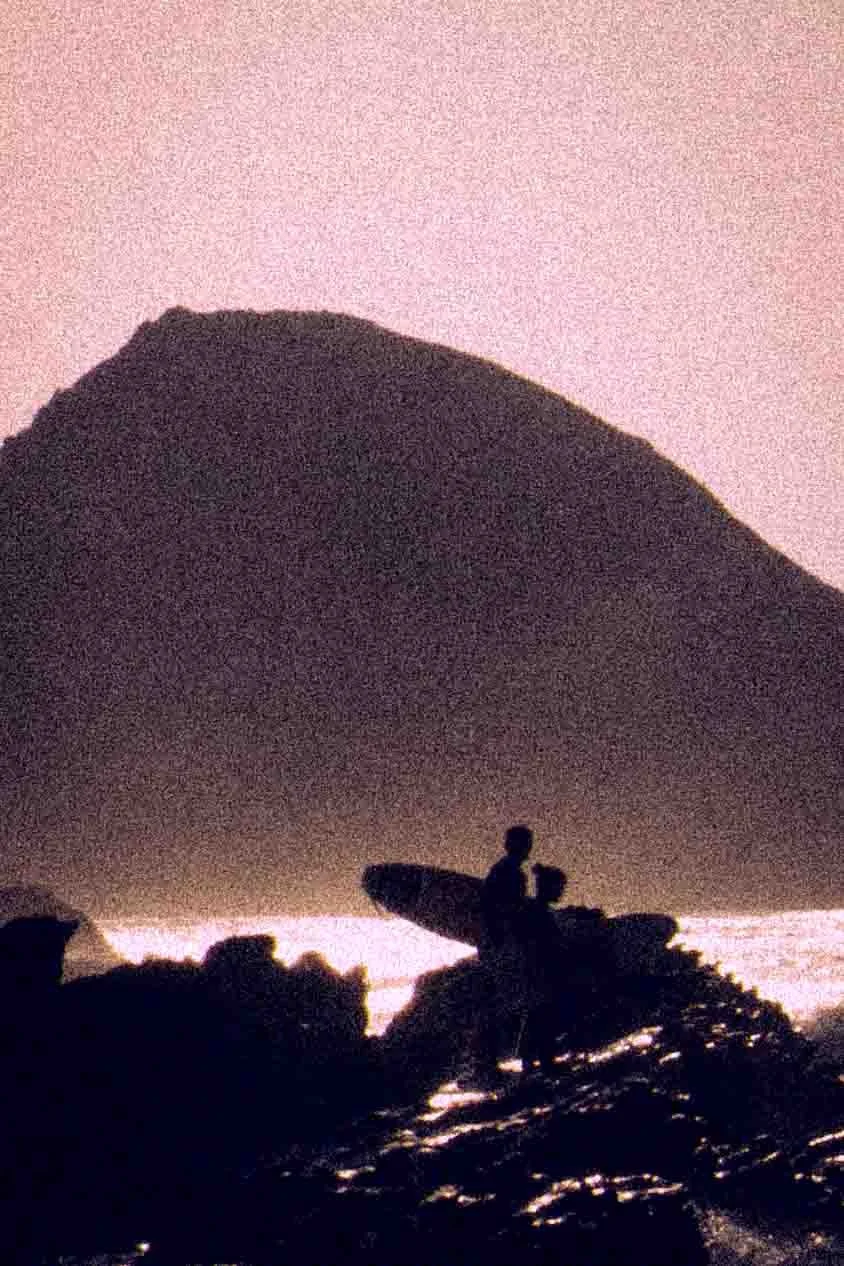 Silhouette of a person standing on a rocky surface, holding a surfboard, with a large mountain or hill in the background during sunset or dawn.