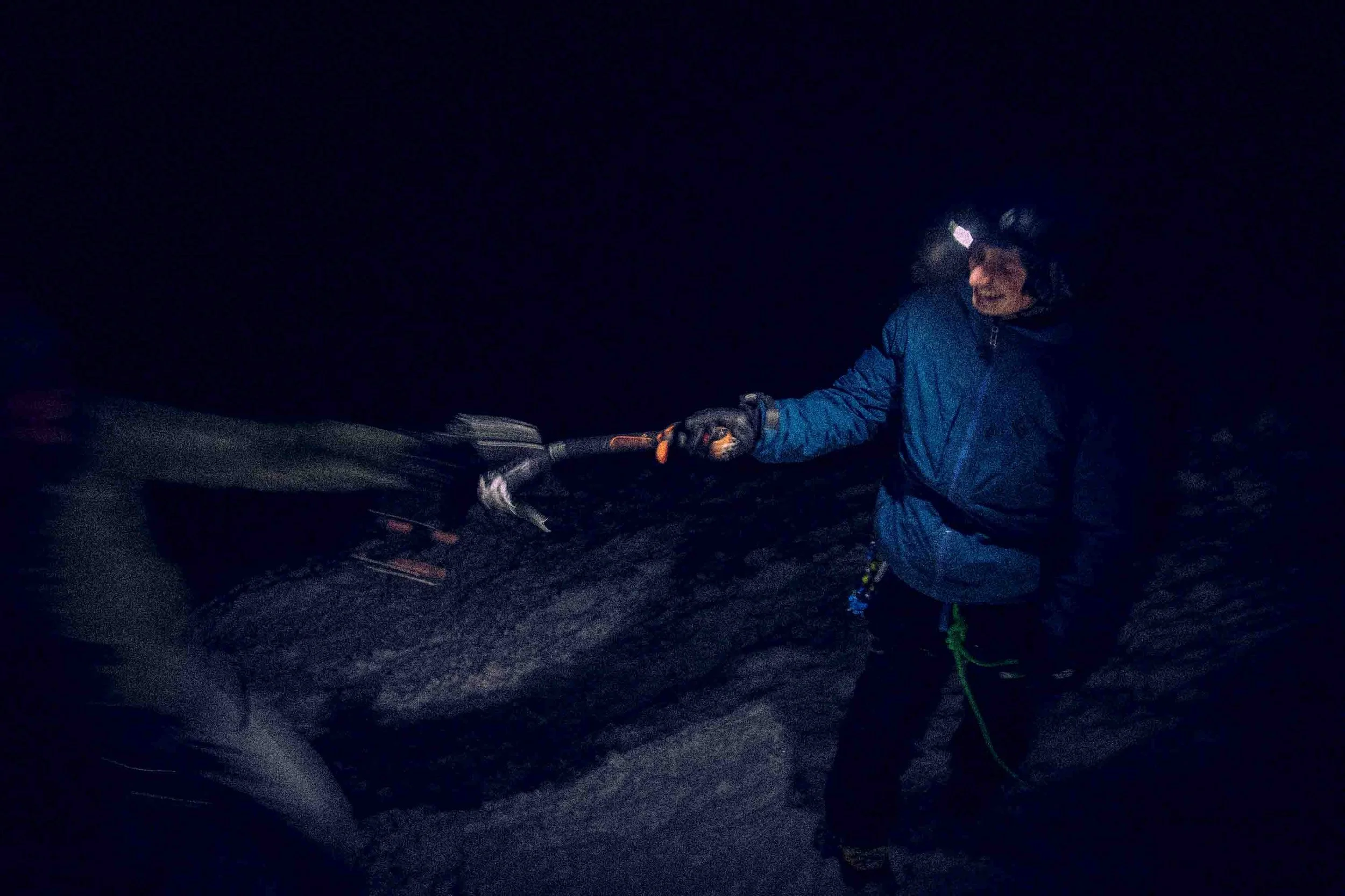 Person ice climbing at night with headlamp, wearing a blue jacket and harness, using an ice axe to chip ice.
