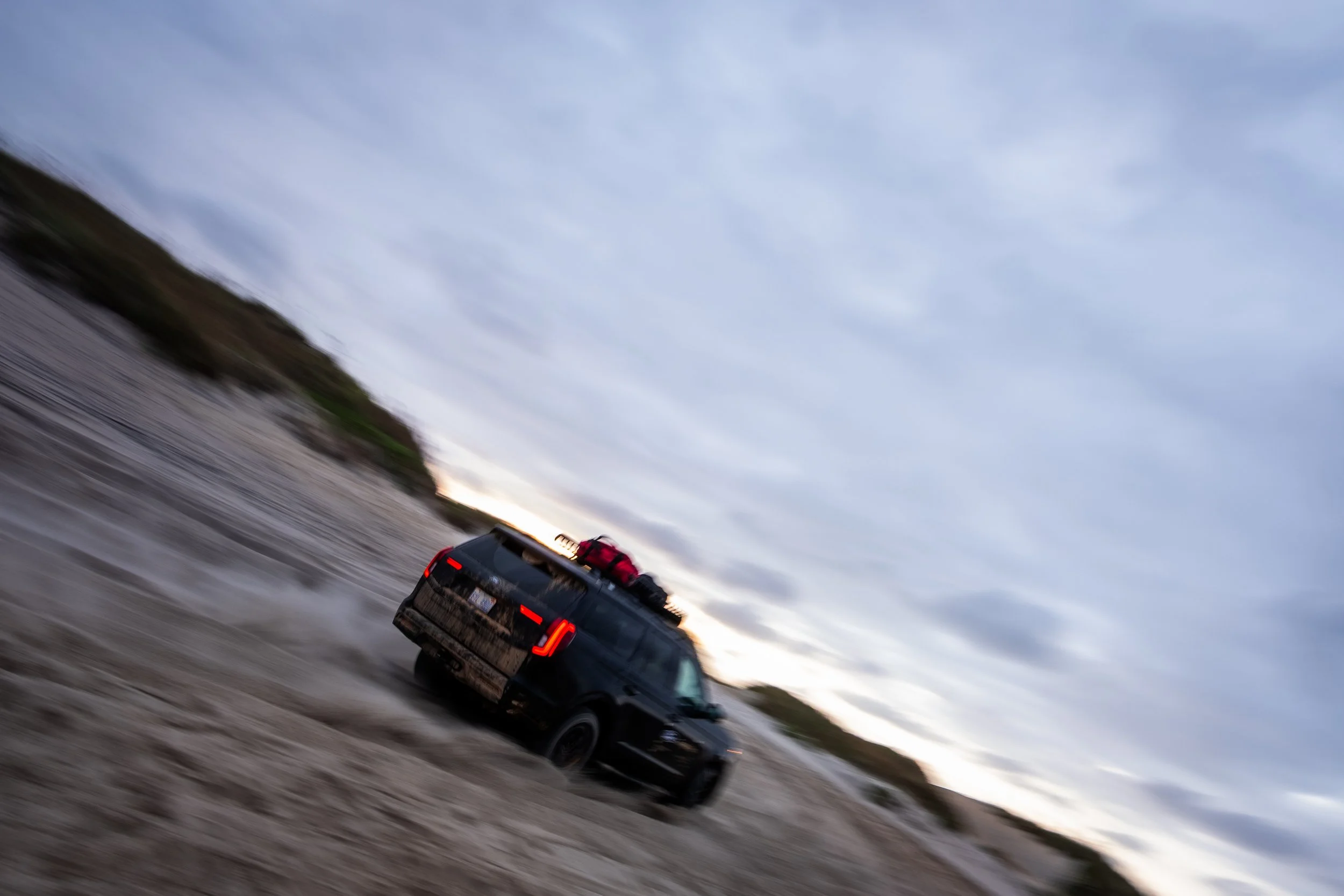 A black SUV driving on a dirt road in a remote, open landscape with cloudy sky overhead, luggage on roof rack.