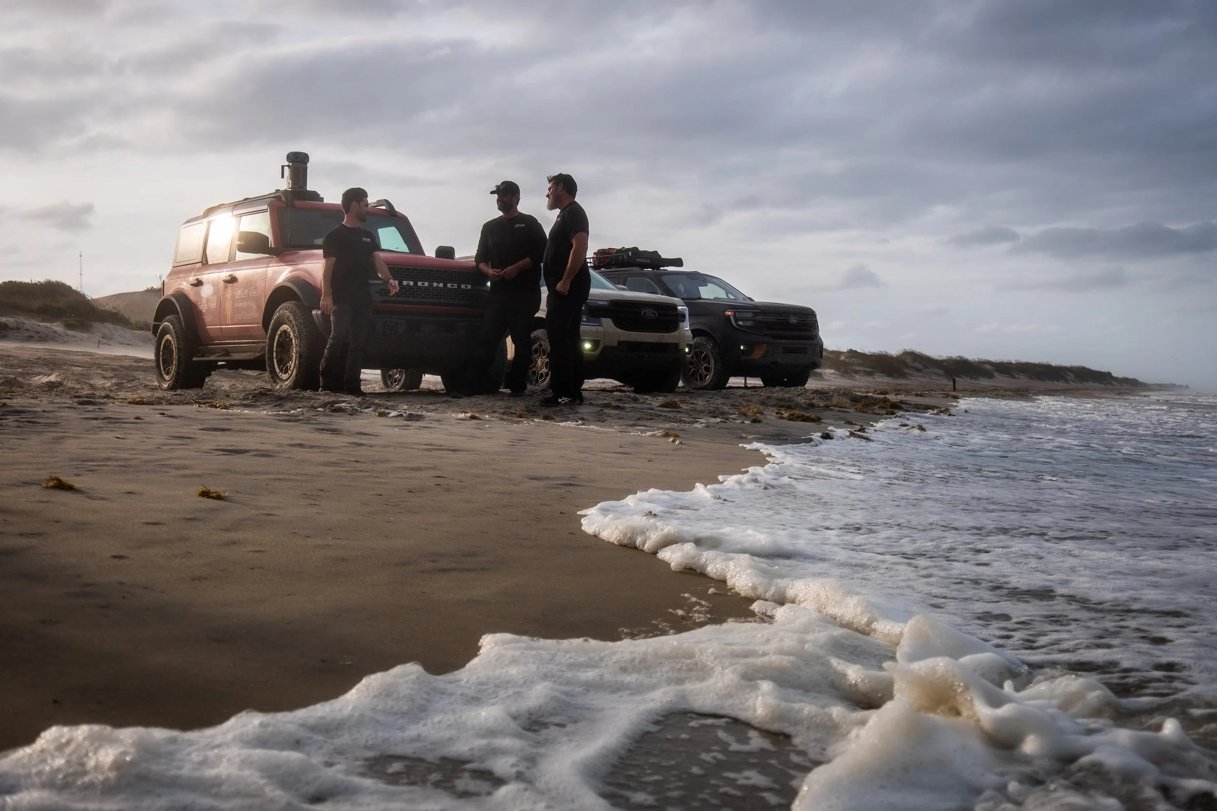 Three men standing next to three off-road vehicles on a beach, with waves in the foreground and cloudy sky overhead.