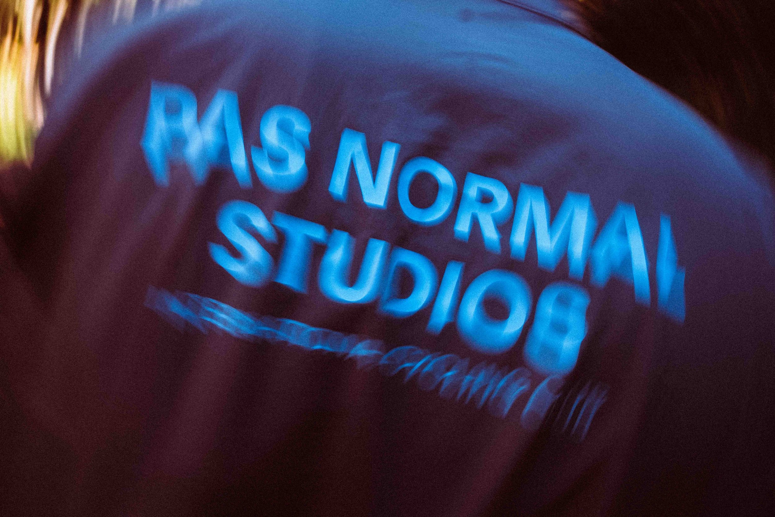 Close-up of a person's face wearing glasses with neon blue text reflecting on the lenses that reads 'PAS NORMAL STUDIOS'.
