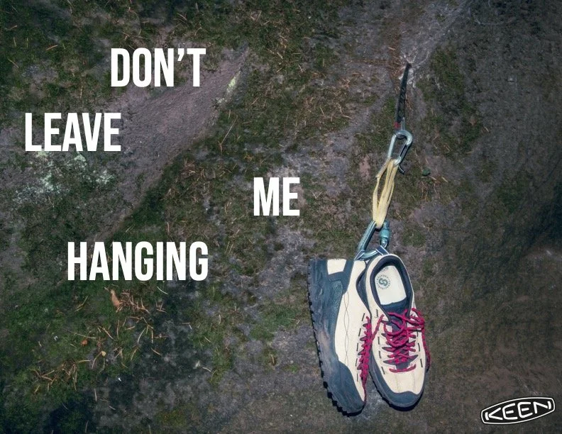 A pair of hiking shoes hanging from a bridge or rock face with climbing gear attached, accompanied by text that says "DON'T LEAVE ME HANGING." Keen photography.