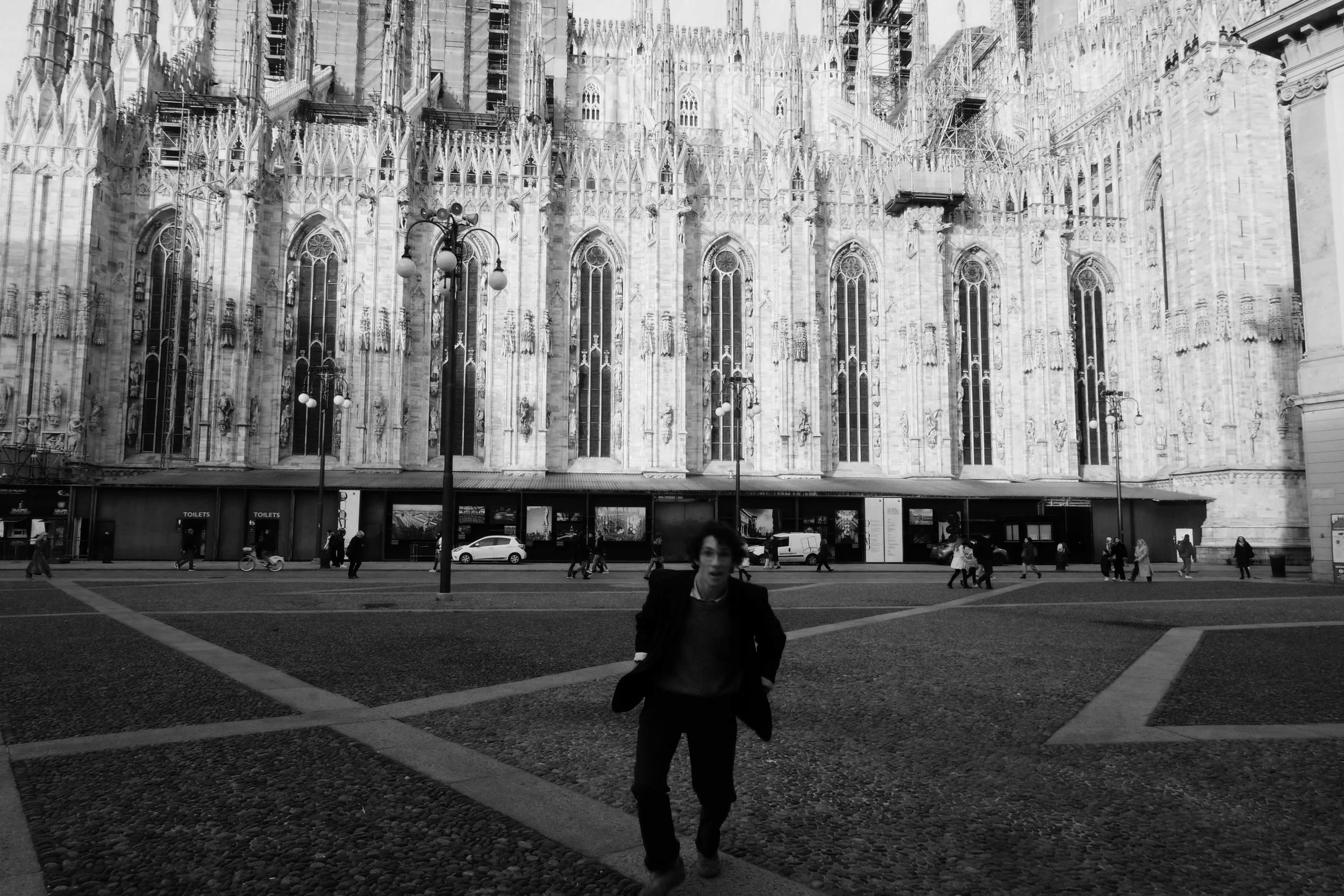 A Black and white photo of a woman walking towards the camera in a large open plaza with the front view of a Gothic cathedral building with tall arched windows and intricate stonework in the background. Several pedestrians are walking along the building and street.