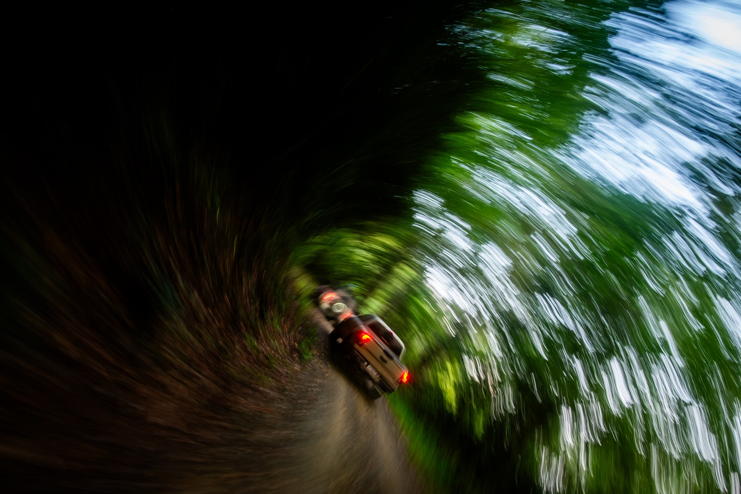 A moving trail of separated cars driving on a narrow, winding dirt road surrounded by dense green trees.