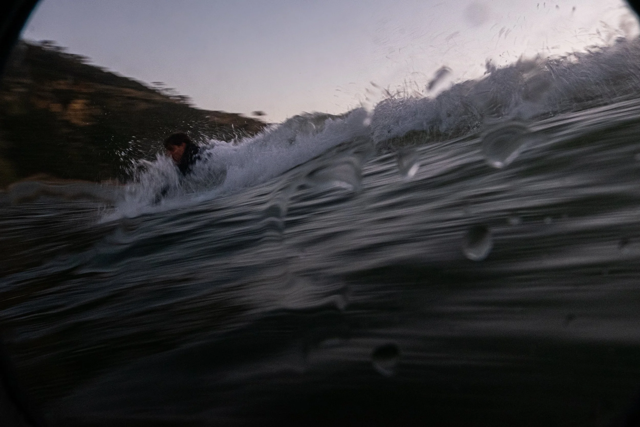A person surfing in the water with a wave in front and a hilly landscape in the background during late evening or early morning.