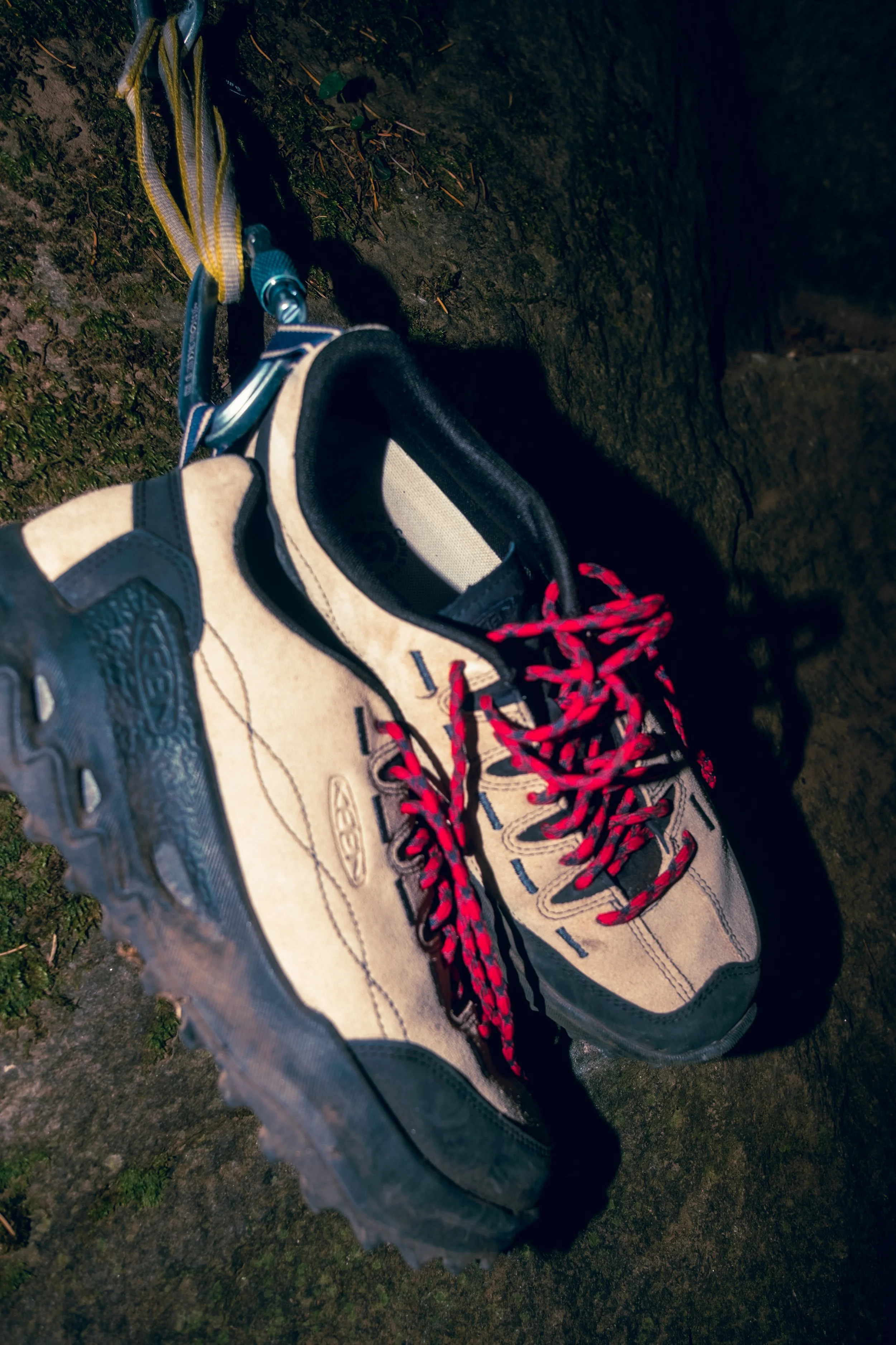 A pair of hiking shoes with red and black laces on a rough, mossy surface, attached to a climbing anchor or metal bolt on a rock wall. Keen photography.