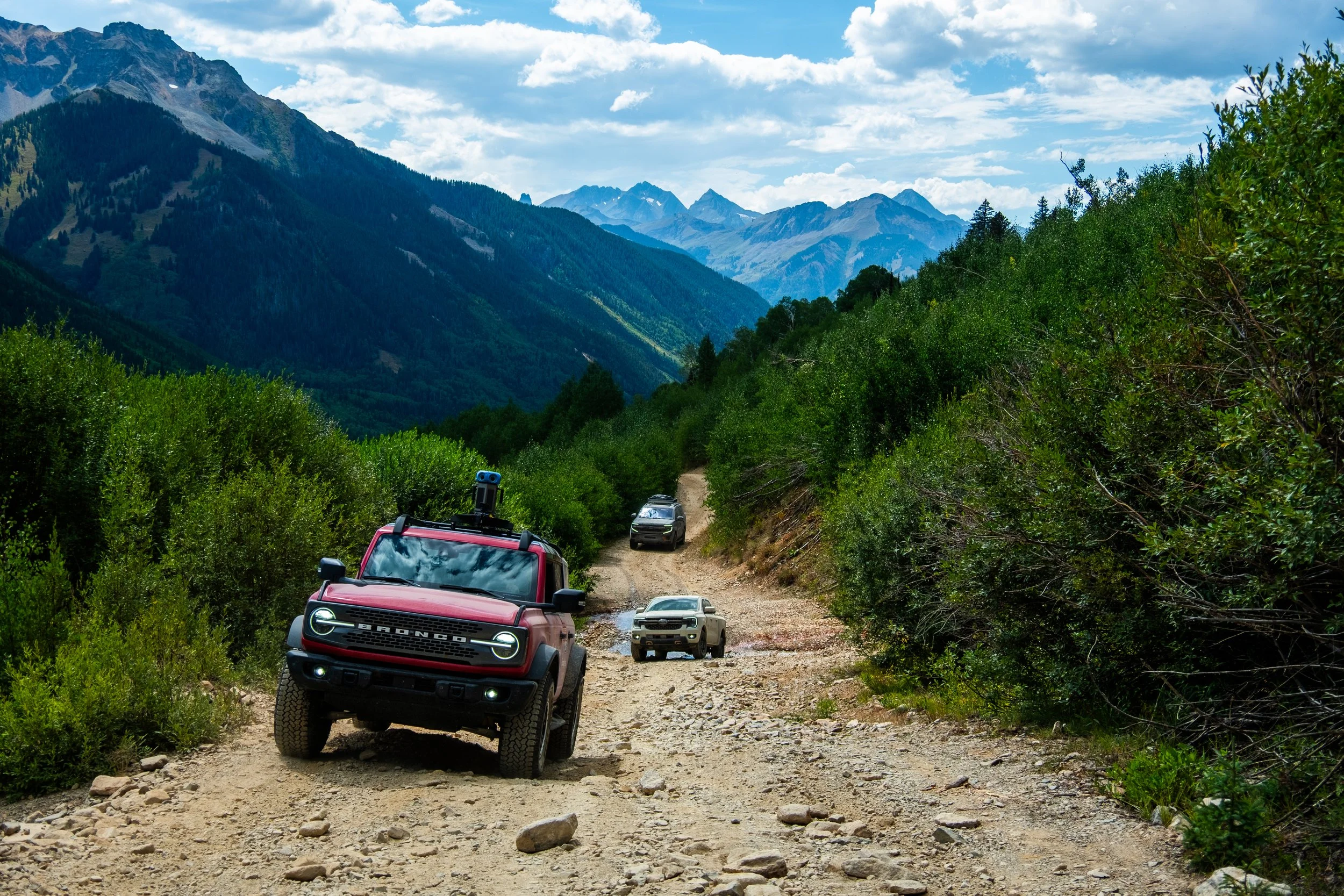 Several off-road vehicles traveling on a rocky dirt trail through a mountainous landscape with green foliage and a partly cloudy sky.