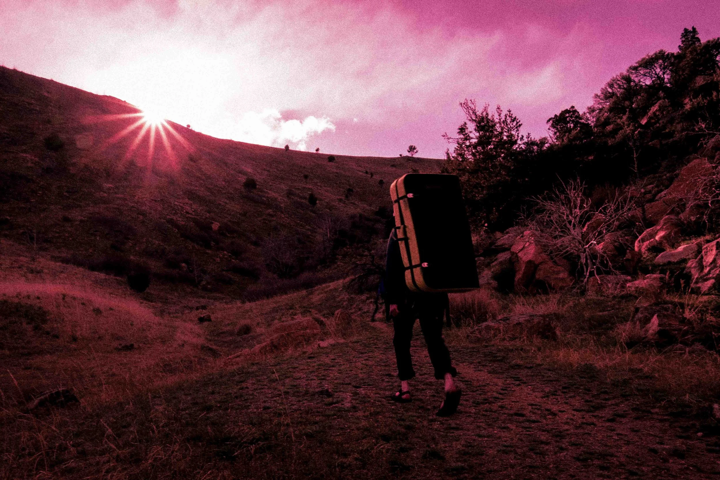 Person walking on a hillside trail carrying a large backpack at sunset with pink and purple sky.