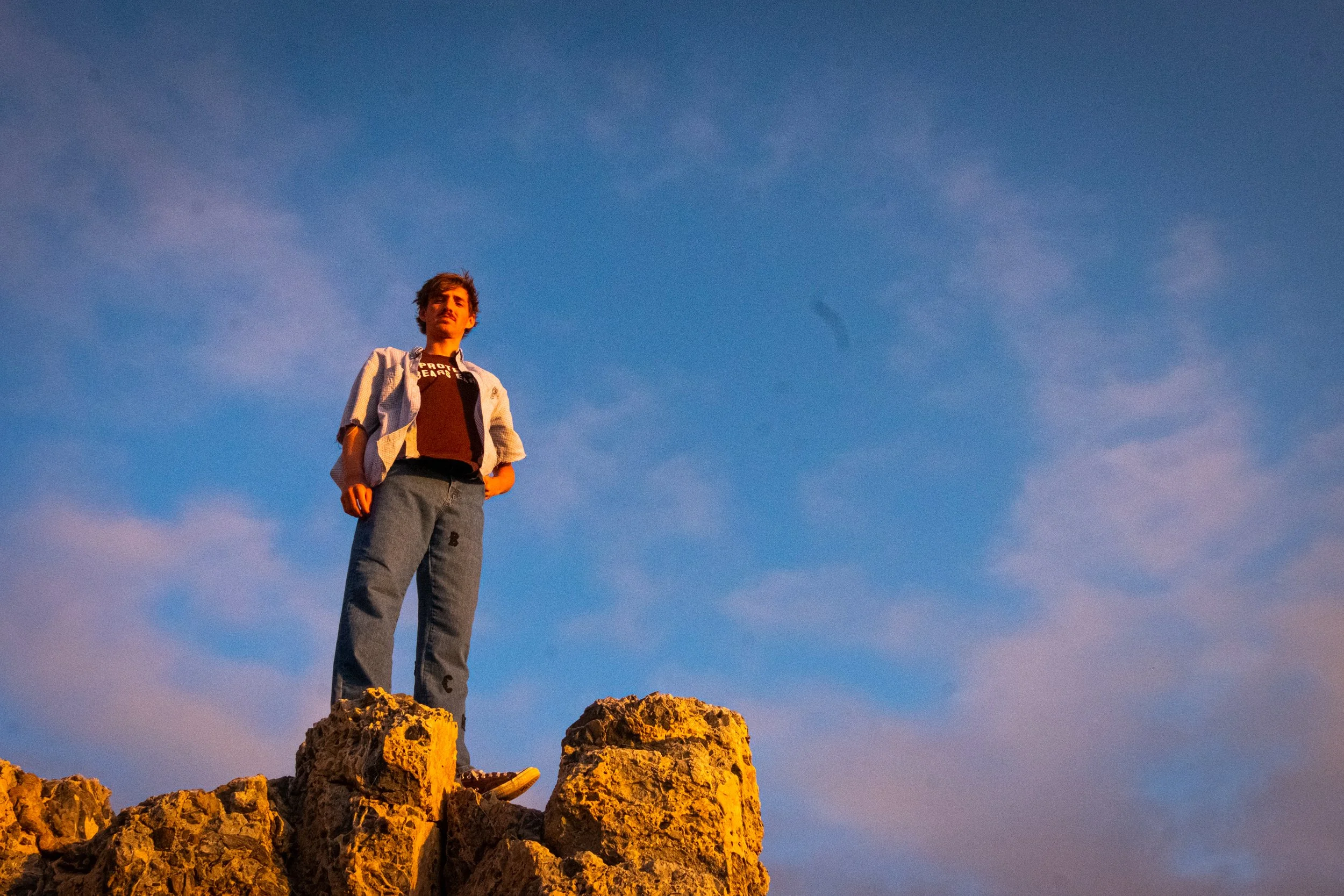 A young man standing on large rocks outdoors during sunset, with a blue sky and some clouds in the background.