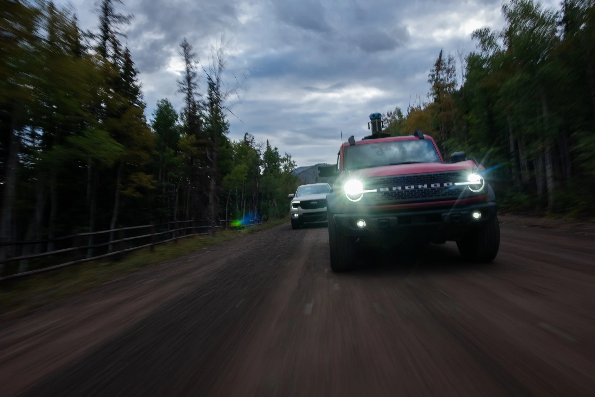 Two cars, a red Ford Bronco and a silver car, driving on a dirt road in a forest with tall trees and cloudy skies, with the Bronco leading.