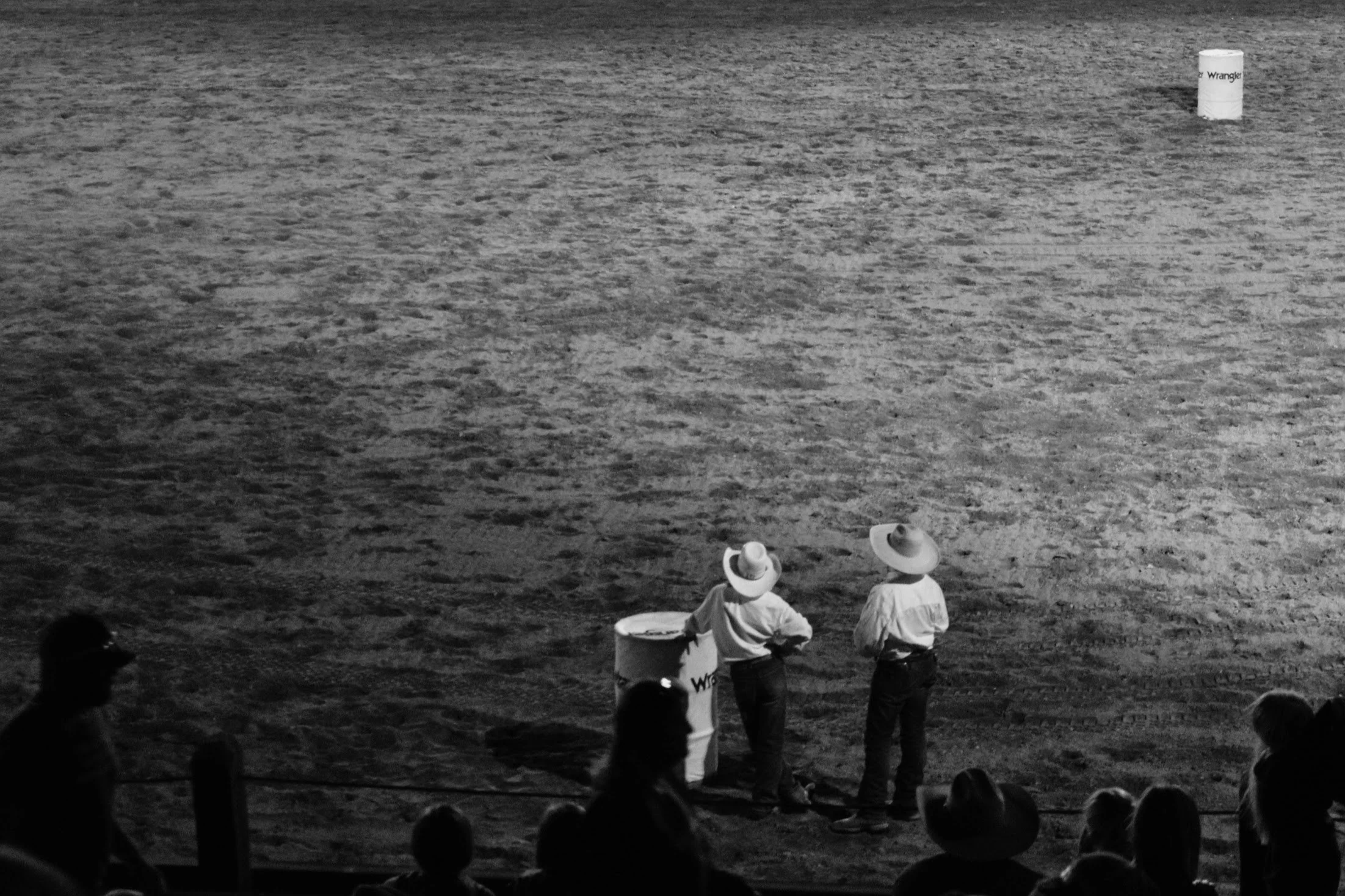 Two cowboys with hats standing on dirt ground near a barrel, with a person sitting and a crowd in the foreground, and a white barrel in the background.