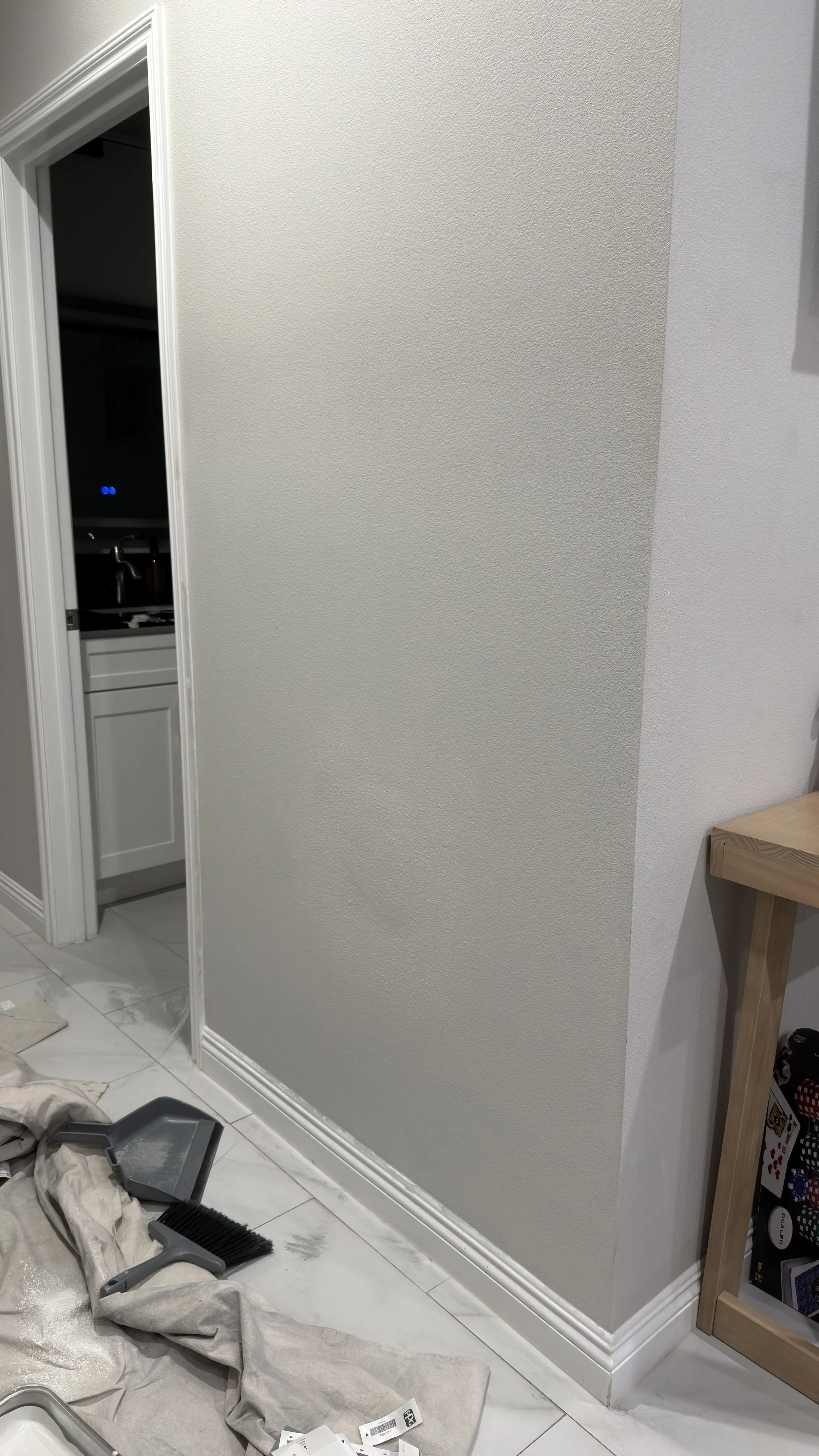 A room corner with renovation tools on the floor, including a dustpan and brush, and a partially painted wall with a baseboard. Part of a kitchen is visible through an open doorway.