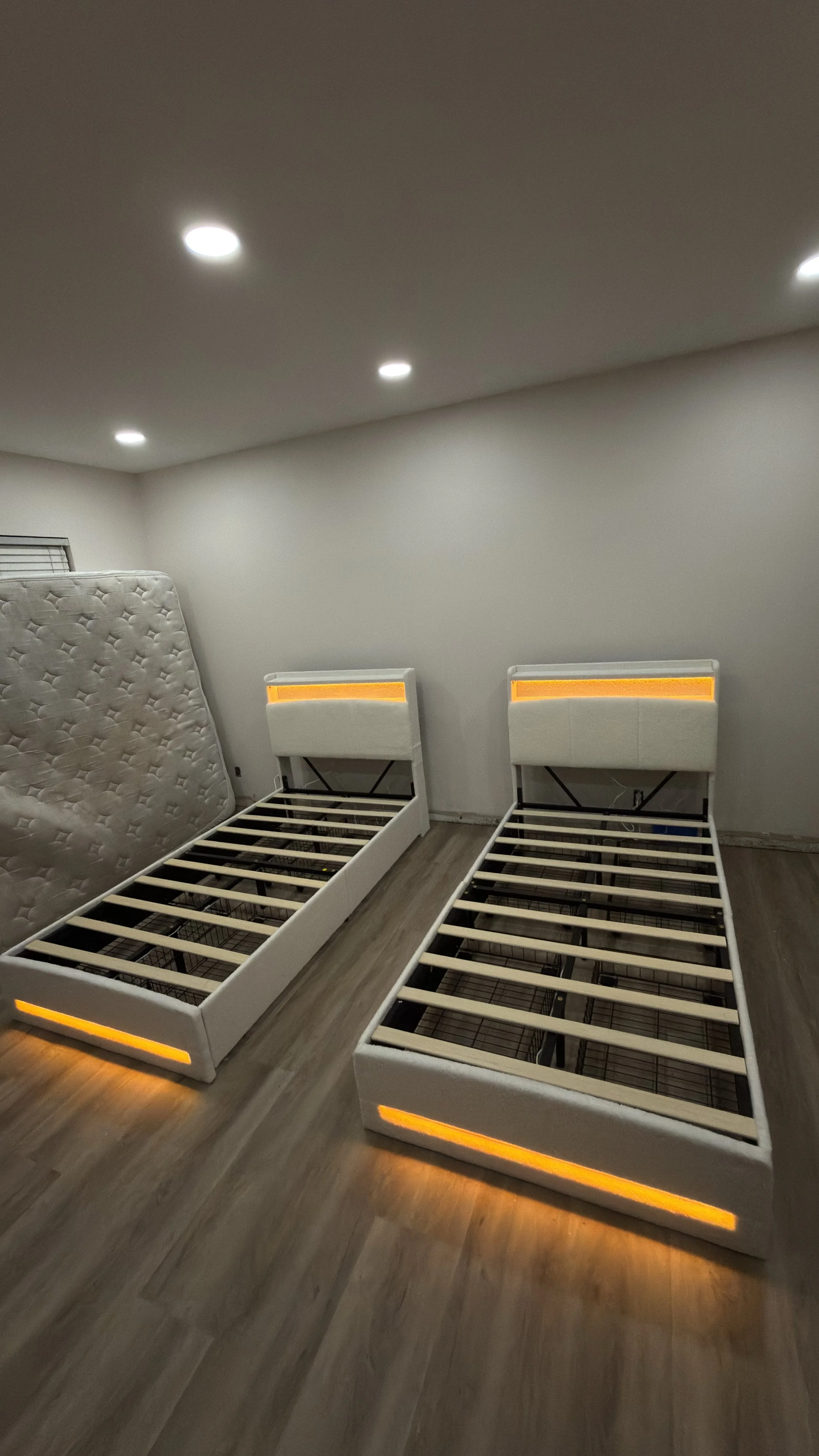 Two bed frames with illuminated headboards and footboards, no mattresses, in a room with wood flooring and a white wall, and a single mattress standing upright against the wall.